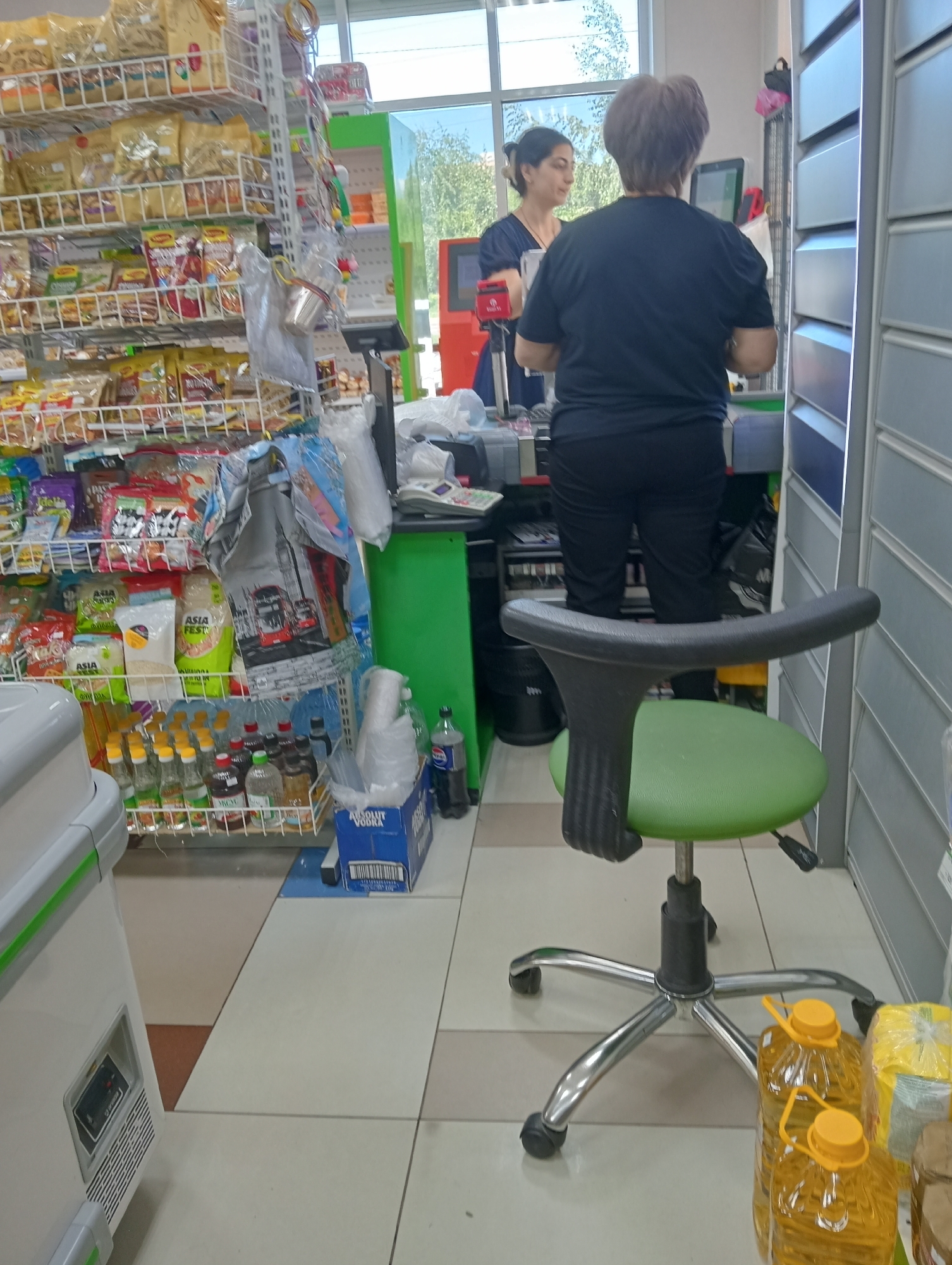 Two women are in what appears to be a convenience store. The woman on the left, wearing a dark blue t-shirt, is looking at a computer screen. The woman on the right, wearing a black t-shirt and black pants, is standing with her back to the camera, also facing the computer screen. In the foreground, a green office chair with a silver base is positioned facing away from the viewer. Shelves stocked with various food items and drinks line the background. To the right, a set of metal cabinets is visible. The floor is tiled in a light color.