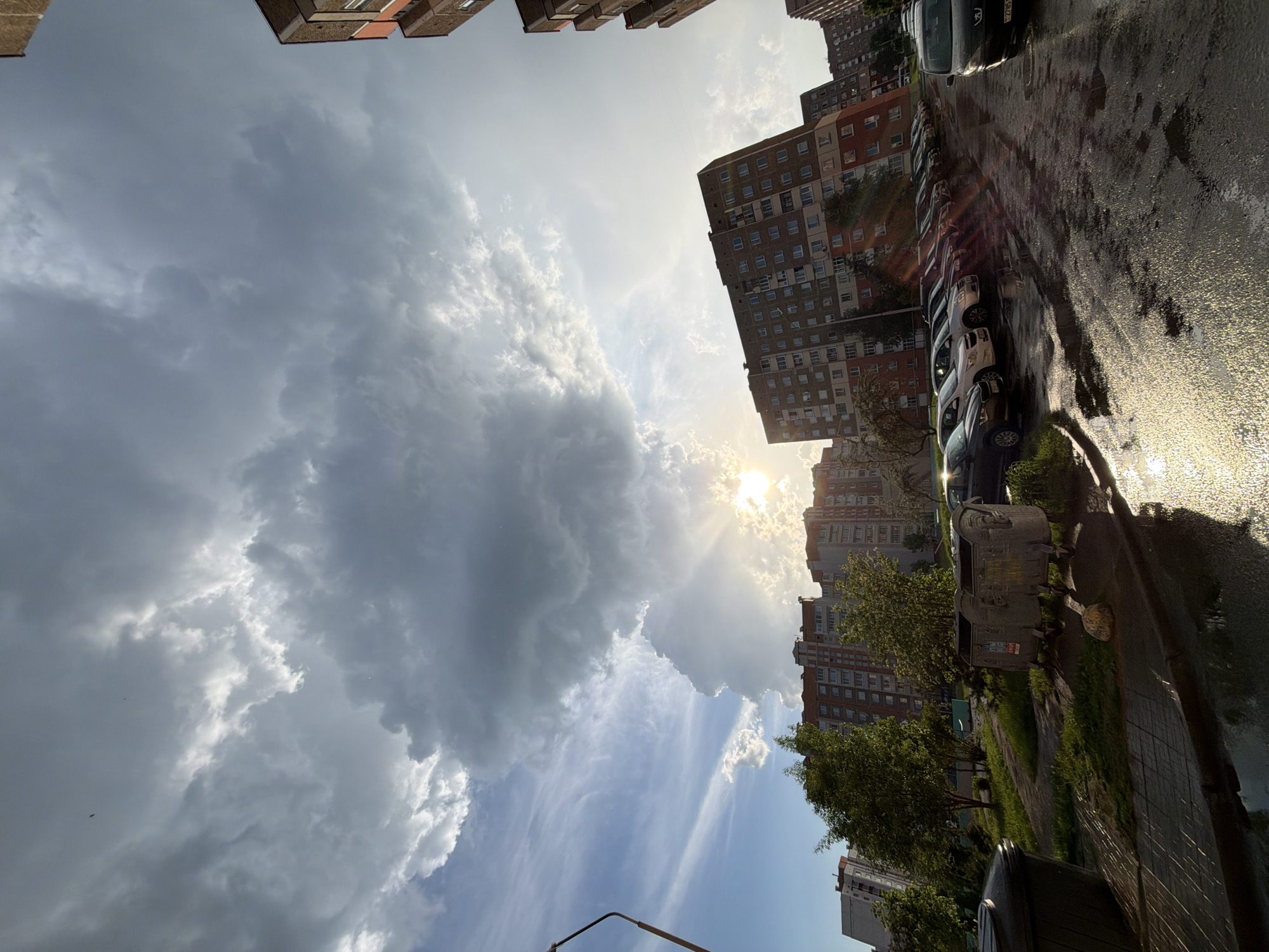 The image depicts a street scene in an urban area after a rain. Tall apartment buildings frame the top and right of the frame. The street is wet, with reflections of the sky and buildings on the ground. Cars are parked along the street, and a trash receptacle is visible on the right side. The sky is filled with dramatic clouds, with the sun shining through them.