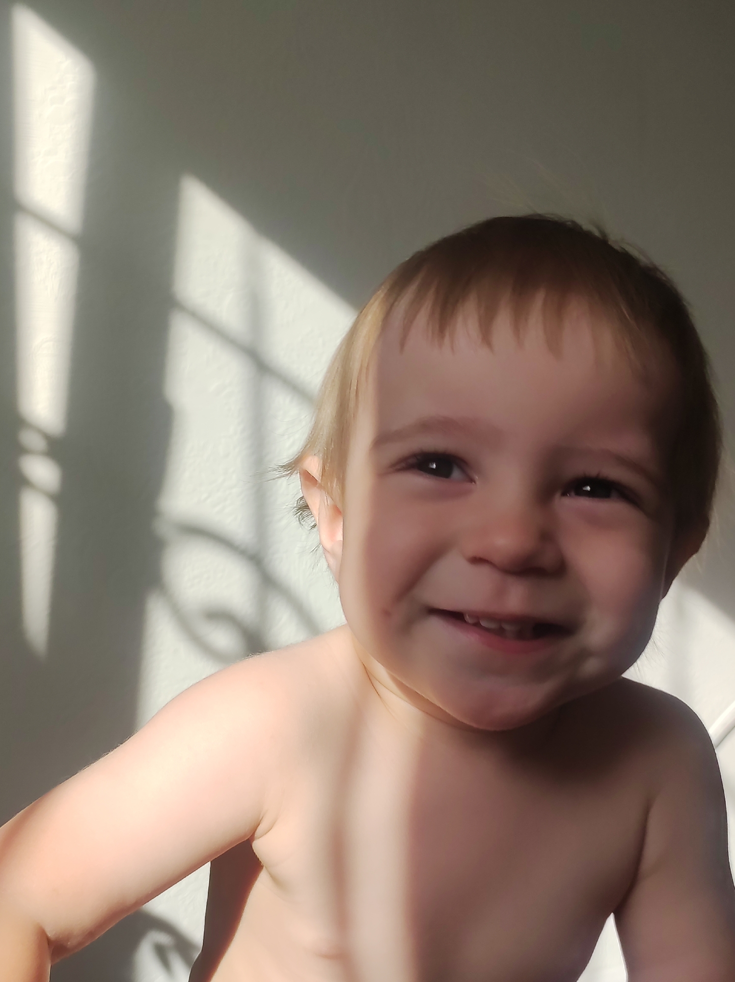 A young child with light skin and blond hair is smiling at the camera. The child appears to be shirtless. The background consists of a white wall with shadows cast by what appears to be a window or a barred structure. No information indicates the location as the city of Chaikovsky, Russia.