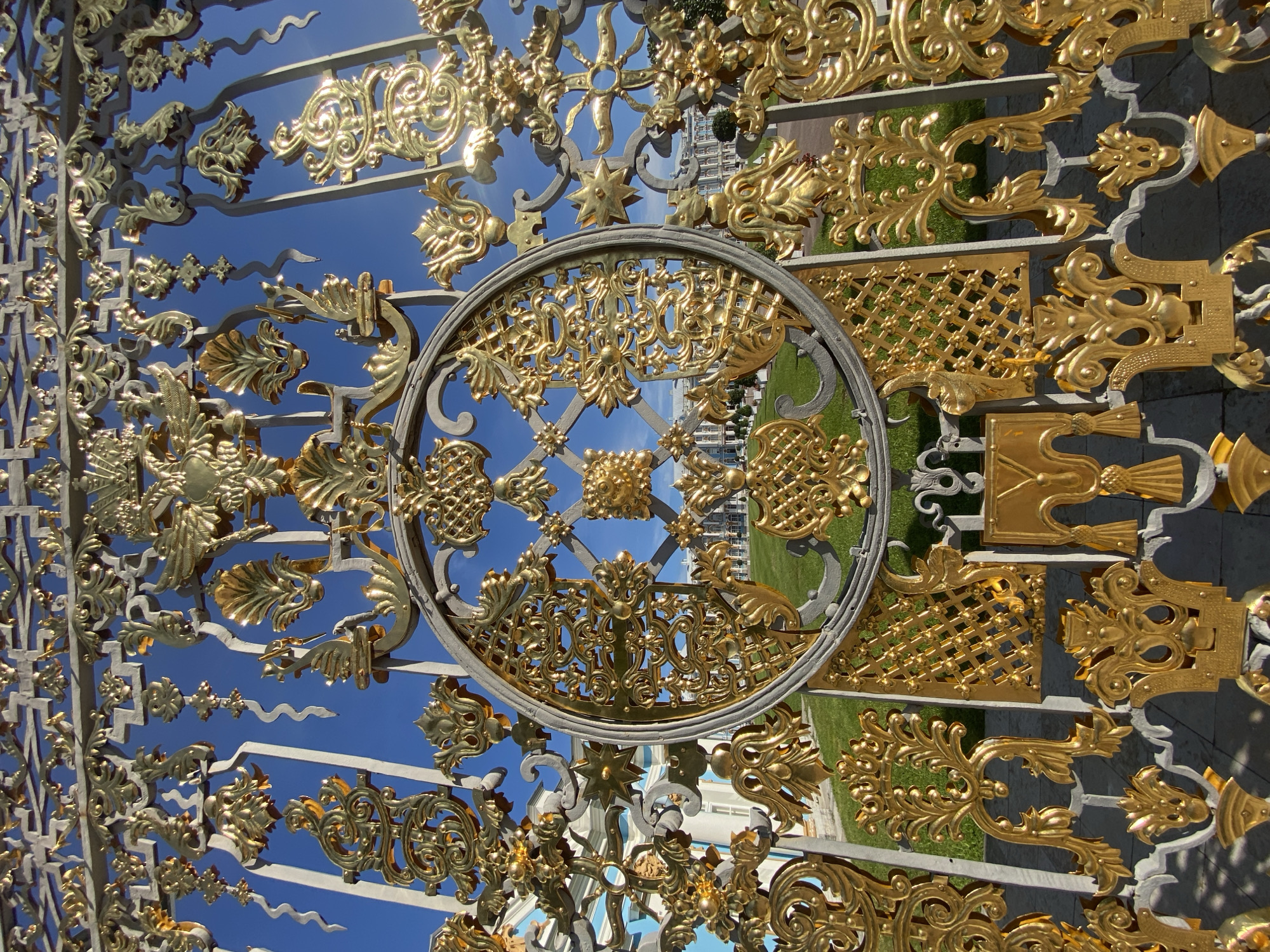 The image shows a close-up of an ornate golden gate, partially obscuring a view of the Catherine Palace in Pushkin, Russia. The gate is made of wrought iron and adorned with intricate gold-plated decorative elements, including floral motifs, swirling tendrils, and geometric patterns. The sun glints off the polished gold, creating bright highlights. Beyond the gate, a section of the Catherine Palace is visible, characterized by its light-colored facade and blue accents. A well-maintained green lawn stretches in front of the palace. The sky above is a clear, vibrant blue. The perspective is from a low angle, looking up through the gate. No people are visible in the frame.