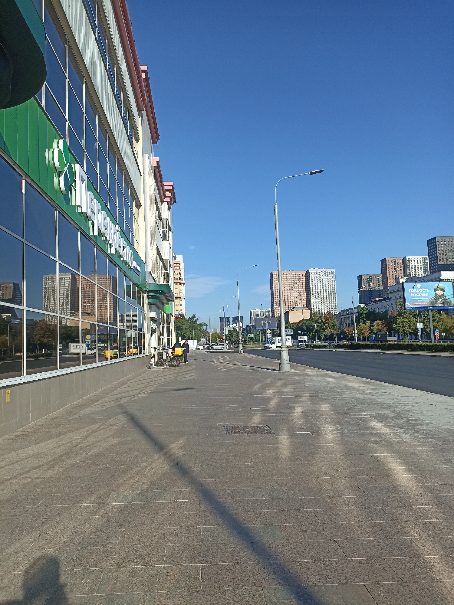 The image depicts a street scene in Moscow, Russia, specifically on a sidewalk adjacent to a modern building with large reflective windows. The building features a green facade with a white clover-like logo and Russian text on its exterior. Across the street, there are several multi-story residential buildings, some of which are a light peach color, others are a mix of white and grey, and some are dark grey. A large billboard is visible on the right side of the street, displaying an image of a soldier with the Russian text "ГОРДОСТЬ РОССИИ!" which translates to "PRIDE OF RUSSIA!". The sky is clear and blue, suggesting a sunny day. The sidewalk is paved with large granite-like tiles and features a shadow of a person cast on it, along with the shadow of a lamppost. Two electric scooters are parked on the sidewalk, and a person wearing a yellow jacket with a black backpack is standing near them. Further down the street, vehicles, including cars and a white truck, are visible. The overall impression is a contemporary urban environment in Moscow.