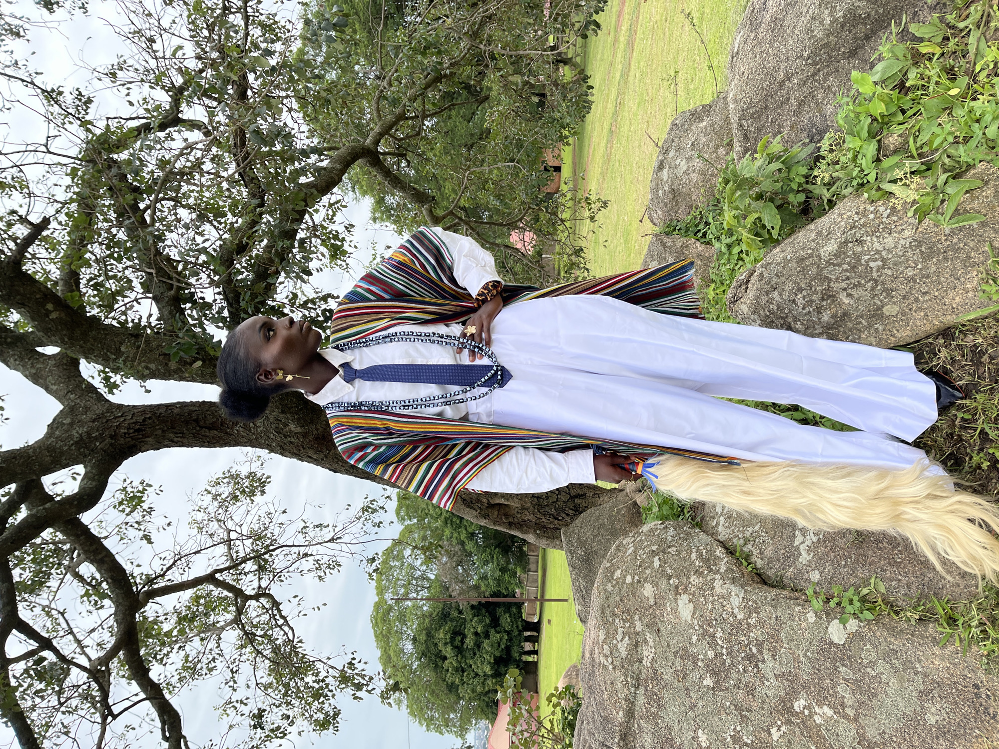 A Black woman stands outside, posing by a large tree and rocks. She is wearing a white shirt, white trousers, and a colorful, striped shawl draped over her shoulders. A blue tie with white patterns is visible around her neck, and she has a gold earring in her visible ear. Her hair is pulled back in a bun. A long, blonde tail-like object, possibly an accessory, hangs from her side.
The background consists of a grassy field with some trees and buildings in the distance. The weather appears overcast, suggesting it could be morning or afternoon. The presence of large rocks and a mature tree indicates a natural outdoor setting, likely in a park or rural area. The woman's posture and gaze suggest a posed photograph, possibly for a cultural or fashion-related purpose.
The location context provided is Jos, Nigeria. There is no visible text in the image.