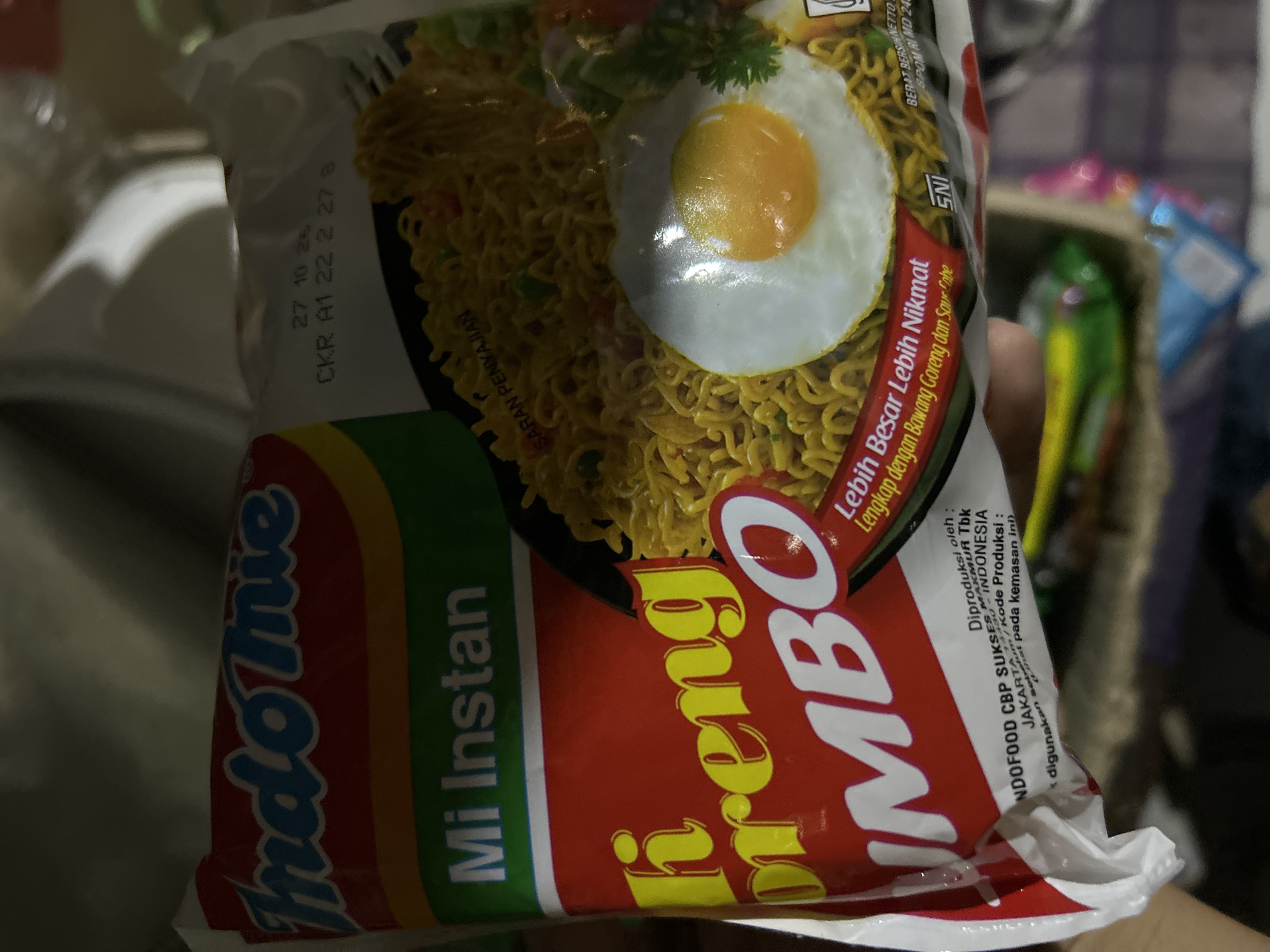 A person is holding a packet of Indomie Mi Goreng Jumbo, a brand of instant noodles, in what appears to be a dimly lit indoor environment, possibly a home or a store. The packet is predominantly red with yellow and green accents. The "Indomie" logo is clearly visible in blue lettering. Text on the packet indicates "Mi Instan" (Instant Noodles) and "Mi Goreng Jumbo" (Jumbo Fried Noodles). A close-up view shows a fried egg and noodles, suggesting the appearance of the prepared dish. A small fork is also visible on the right side. Text at the bottom provides production details, including "Diproduksi oleh: INDOFOOD CBP SUKSES MAKMUR Tbk JAKARTA - INDONESIA". There is no information in the image that specifically places the scene in Depok, Indonesia.