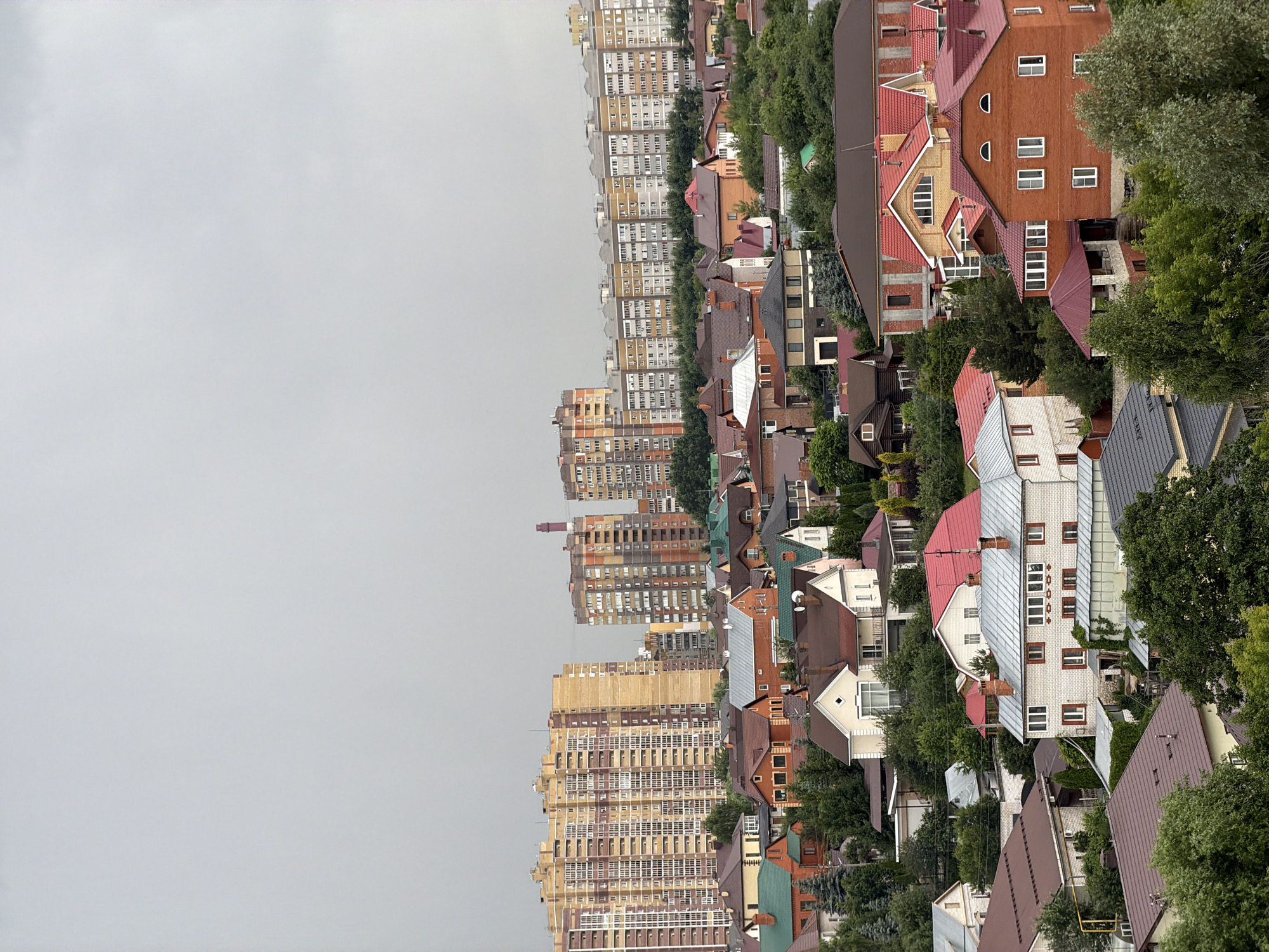 This is a high-angle, eye-level, medium shot of a residential neighborhood under a cloudy sky. In the background, several large, modern apartment buildings are visible, characterized by their multi-story structures and numerous windows. In the mid-ground and foreground, a dense collection of houses of various sizes and architectural styles fills the scene. These houses feature diverse roof designs, many with red or brown shingles, and some have visible chimneys. Trees and green foliage are interspersed throughout the neighborhood, with some trees appearing to be deciduous and others evergreen. The overall impression is of a developed urban or suburban area with a mix of high-rise apartments and detached homes.