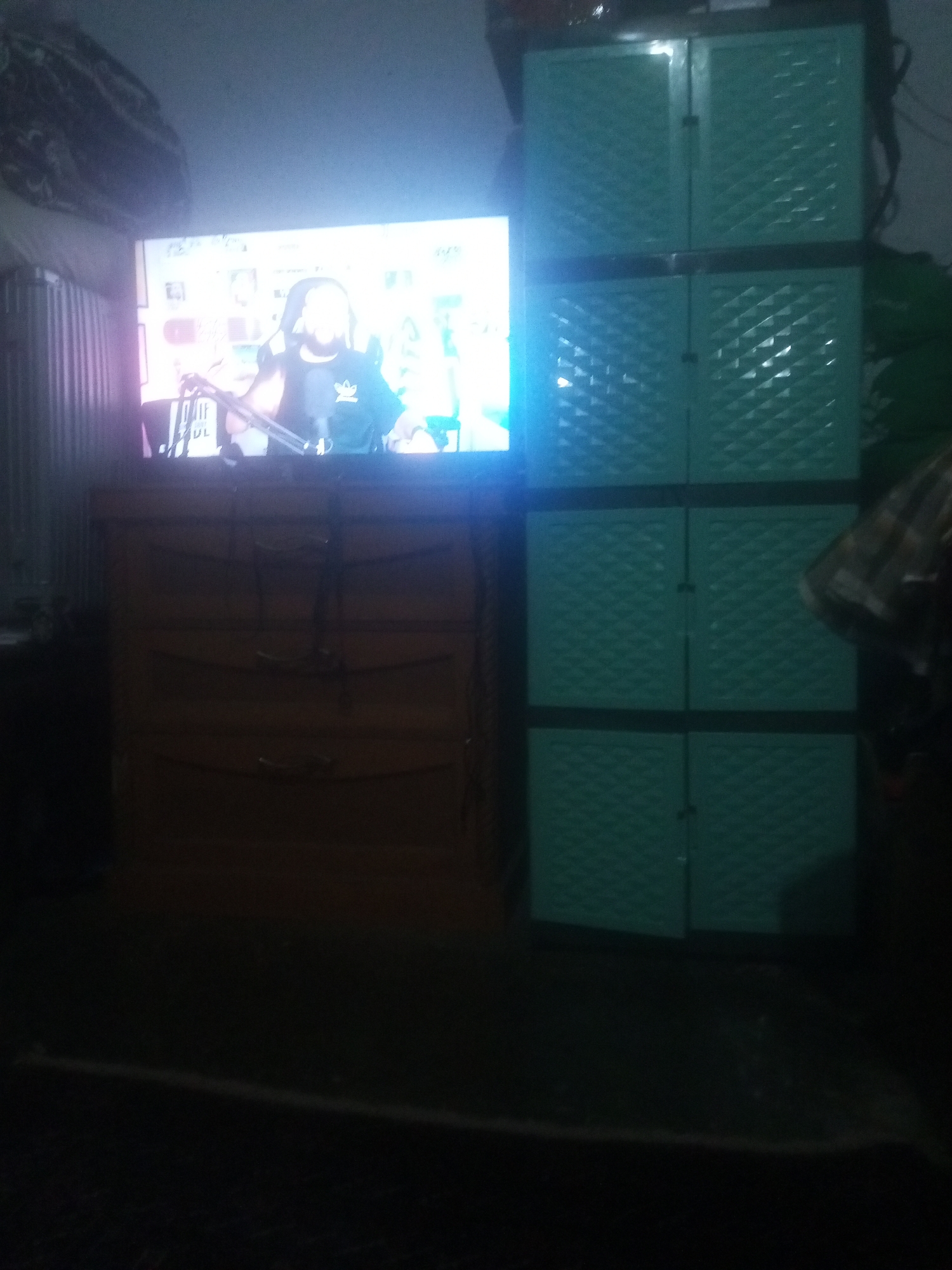 In the city of Tanjungpura, Indonesia, a television screen displays a person in a gaming chair, holding a microphone. To the right of the television, a tall, teal-colored cabinet with diamond-patterned doors is visible. In front of the television is a wooden dresser with multiple drawers. The overall lighting in the room is dim.