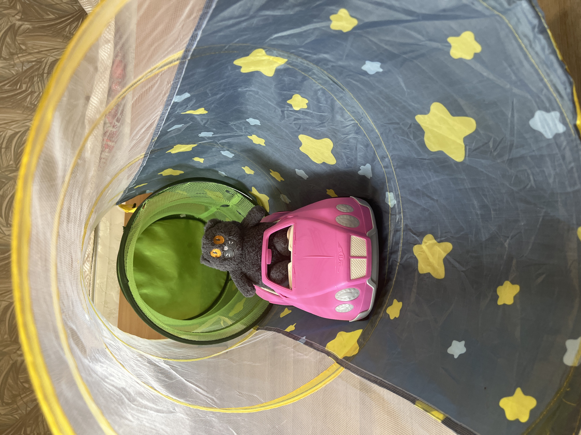 A stuffed grey cat with orange eyes is seated in a pink toy car. The car is positioned within a fabric tunnel that is dark blue with yellow and light blue stars. The tunnel has a mesh section with a green interior at the far end, and a yellow trim around the entrance. The scene appears to be indoors, with a textured wallpaper visible in the background. The image does not provide any information to determine its location in the city of Yekaterinburg, Russia.