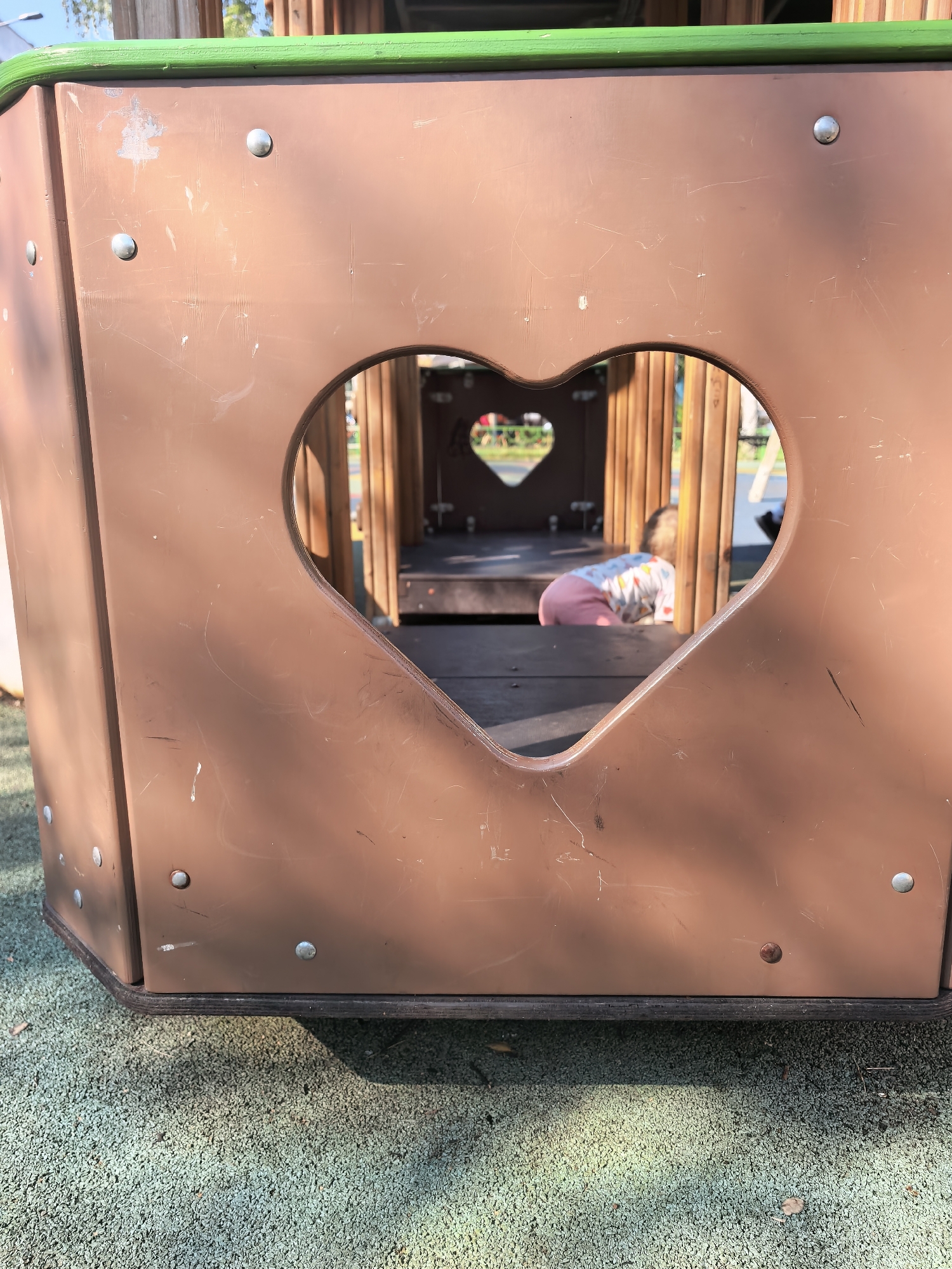 The image shows a close-up of a brown, heart-shaped cutout on a playground structure. Through the cutout, a child wearing pink pants and a patterned shirt is visible crawling on a wooden platform inside the structure. The playground is located on a green, textured surface. The surrounding area is out of focus, but glimpses of trees and sunlight suggest an outdoor setting. The overall scene is consistent with a public playground in Sterlitamak, Russia.