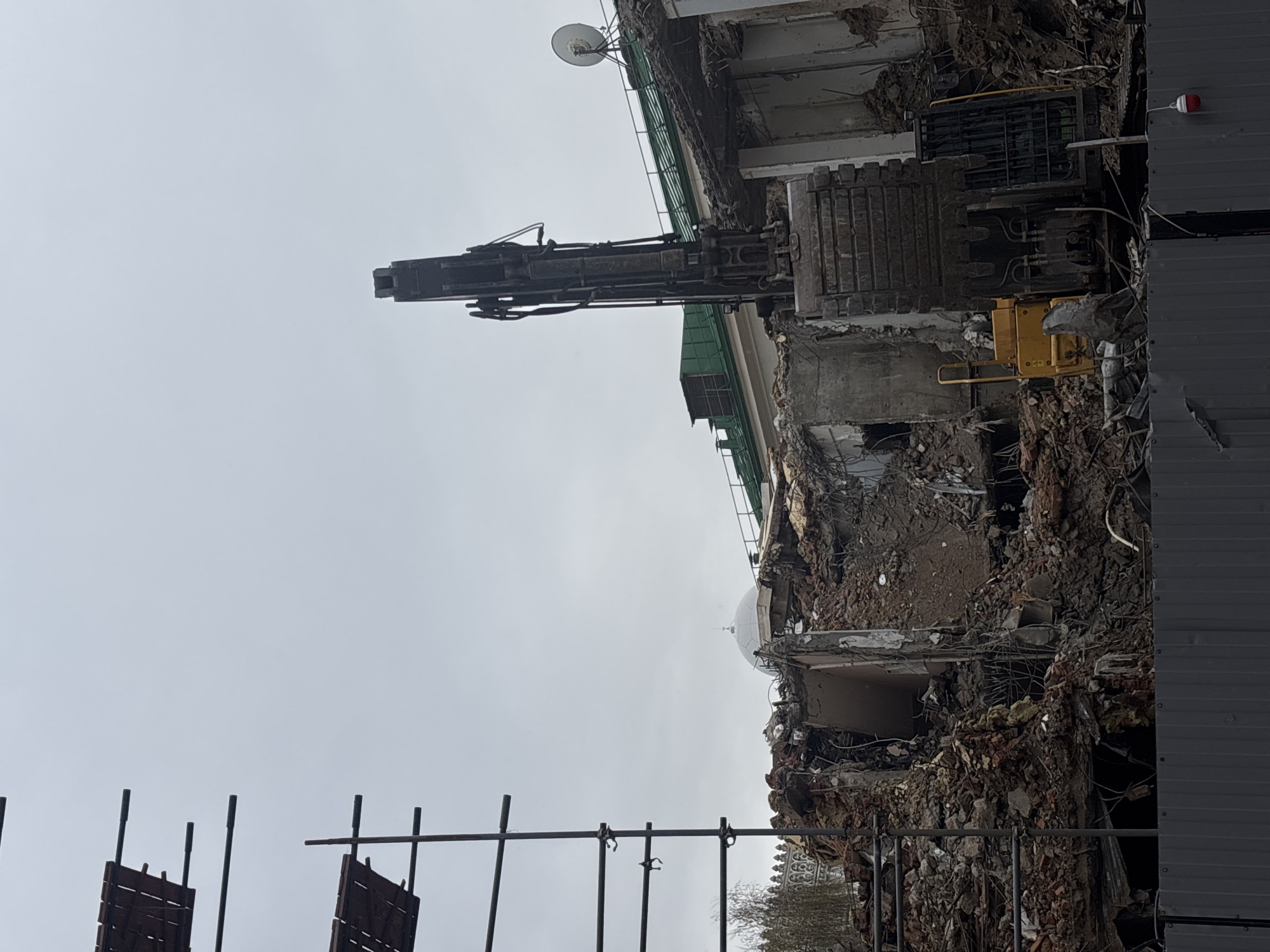 The image shows a demolition site in Moscow, Russia. A large building is partially destroyed, with rubble scattered around. A demolition excavator with a long boom is positioned on top of the remaining structure. Scaffolding and a corrugated metal fence are visible in the foreground. A satellite dish is attached to the building. The sky is overcast.