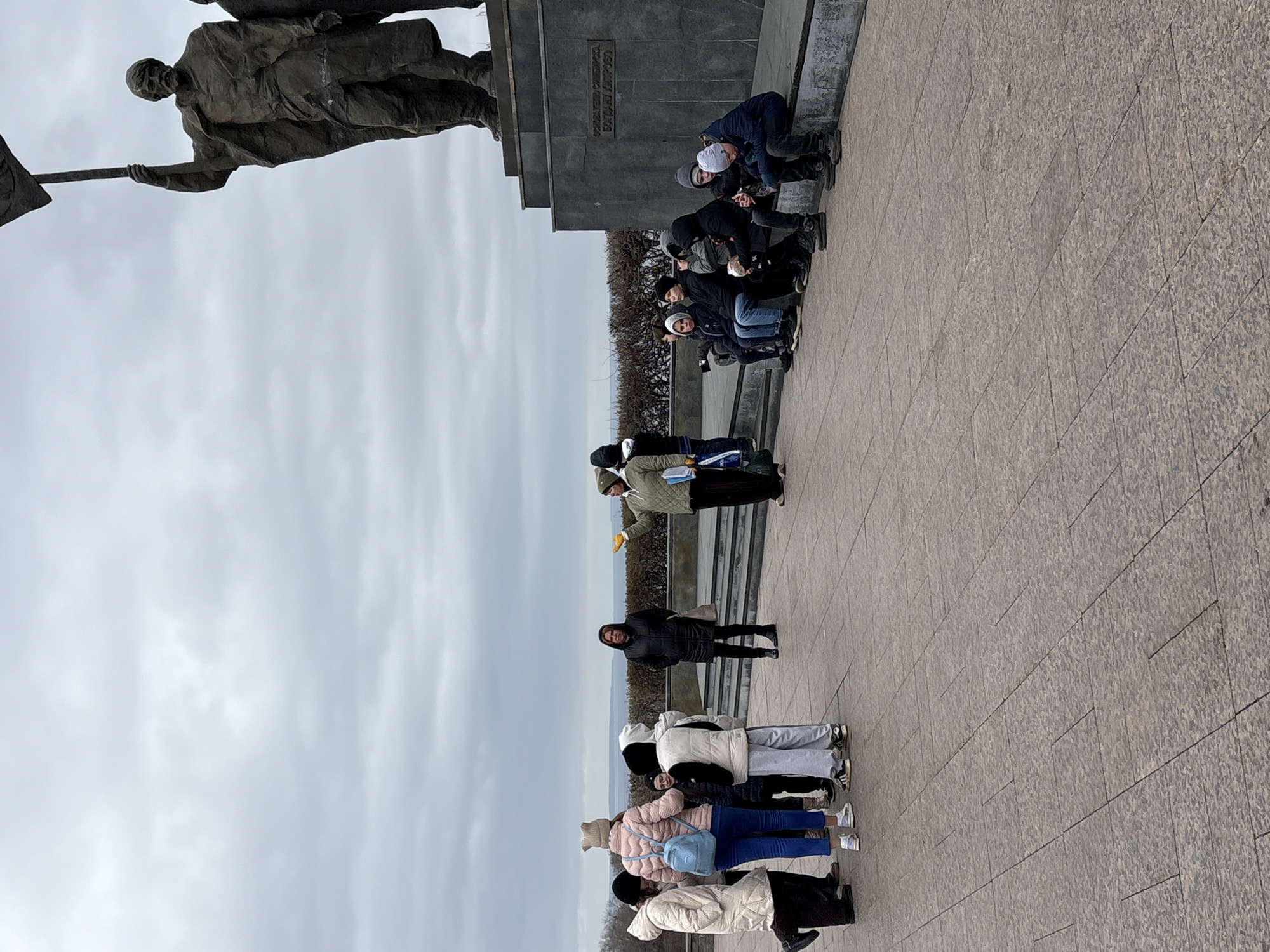A group of people is standing in front of a monument in Ulyanovsk, Russia. The monument is a statue of a man holding a flag. The group is made up of people of different ages and genders. Some people stand on the walkway while others are on the top level near the monument. The environment is cloudy and the ground is paved with gray stone.