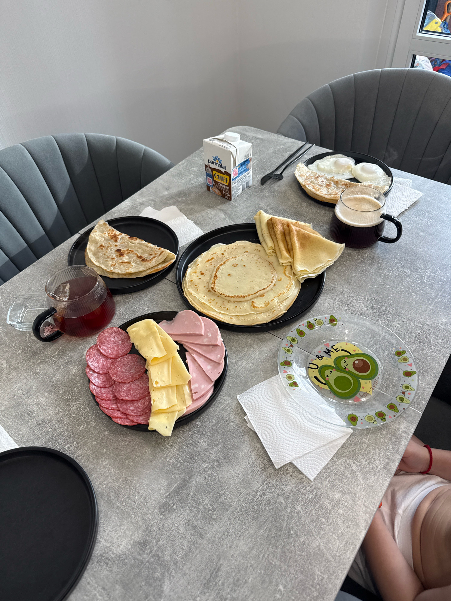 The image depicts a breakfast scene laid out on a grey, textured table. On the table, there are two black plates holding various food items. One plate contains folded crepes, and next to it is a pile of crepes. Another plate displays a cheese and meat platter with sliced salami, cheese, and ham. A glass jug filled with dark liquid, likely tea, sits nearby. A carton of "Parmalat Chef" cream and a mug of dark coffee are also present. A clear glass plate with avocado illustrations and the text "U & ME" is visible. Two grey chairs are partially visible. In the lower right corner, a child's arm, wearing a red bracelet, is visible. The overall impression is that of a home-cooked meal. There is no information to indicate the location in Lyubertsy, Russia, or any specific city.