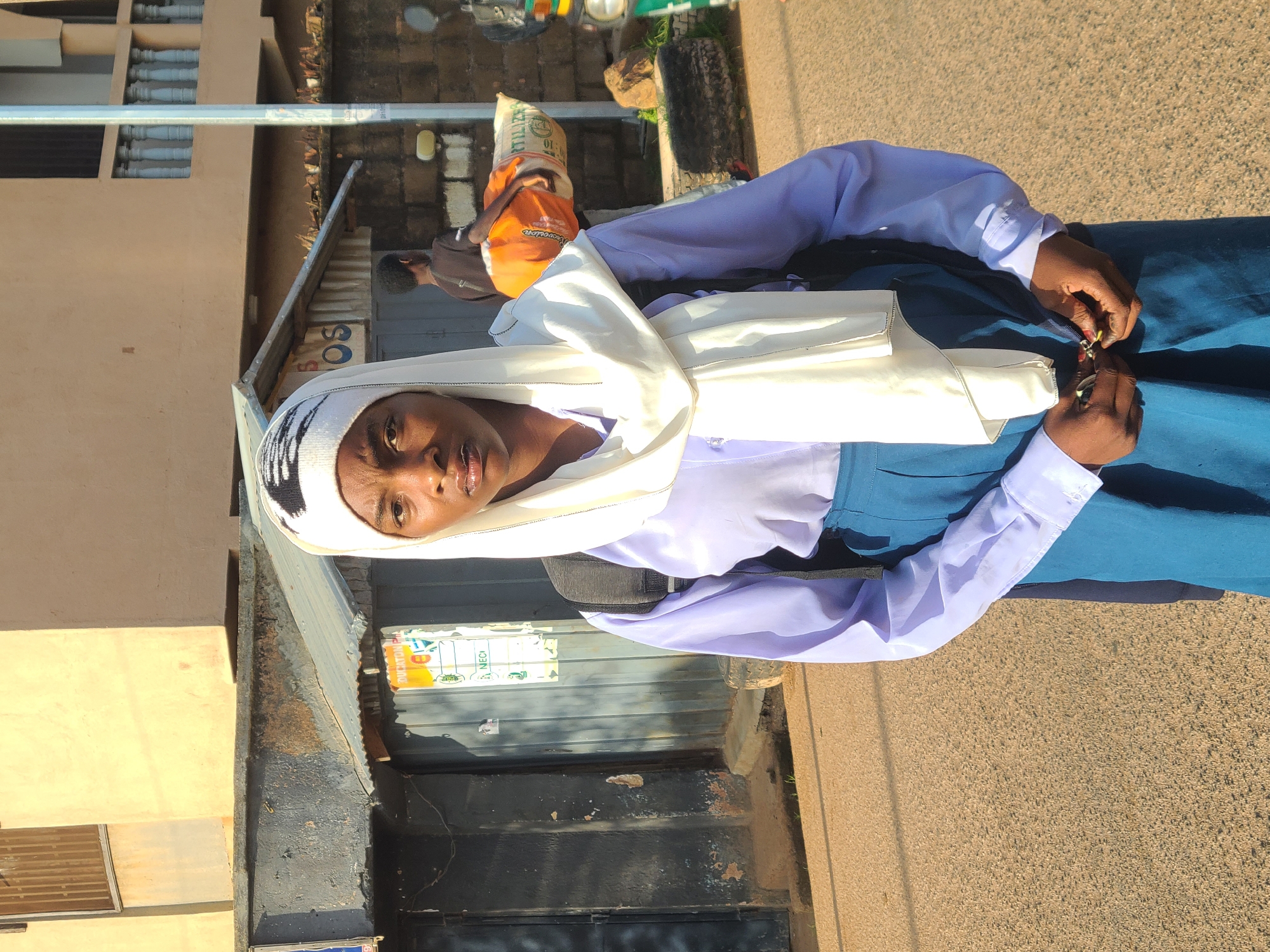 A young woman wearing a white hijab and a light purple long-sleeved shirt with a dark blue skirt stands in front of a building with a corrugated metal roof and a dark gray metal door. She is looking directly at the camera with a neutral expression. Her hands are clasped in front of her waist. A black backpack is visible on her shoulders. In the background, a man carries a large orange and white sack over his shoulder, and part of a motorcycle is visible on the right side of the frame. The ground is paved with asphalt. This scene is located in Jos, Nigeria.