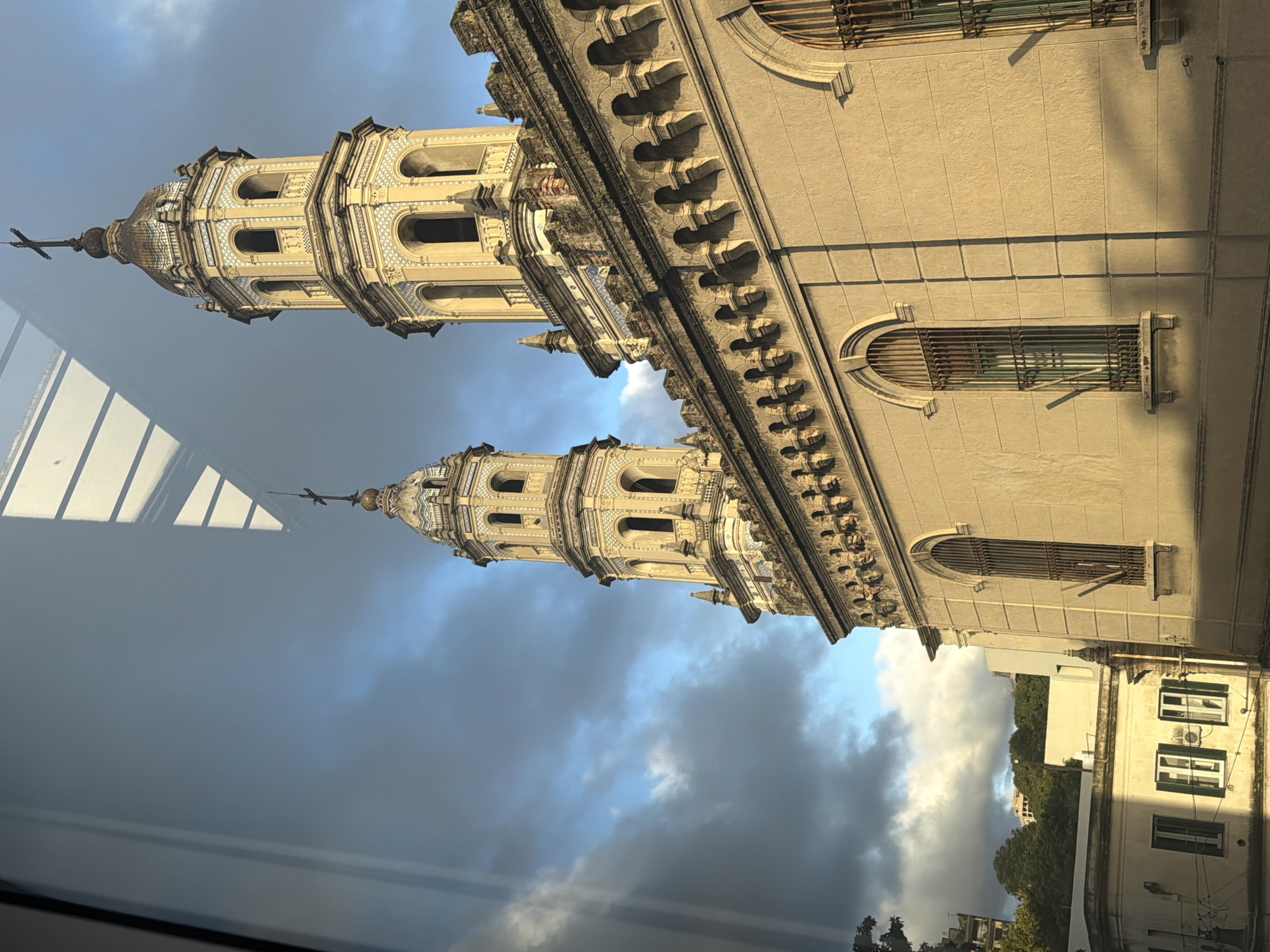 The image captures a grand, ornate building, likely a church or cathedral, featuring two prominent bell towers. The architecture is characterized by a light, warm-toned facade adorned with intricate detailing, including decorative cornices, arched windows with metal grilles, and multi-tiered towers. The towers exhibit beautiful white and blue patterned tiling, open arched sections, and domed tops, each surmounted by a cross. One tower is clearly visible, while the second stands slightly behind it, partially obscured but still reflecting its elaborate design.
The photograph appears to have been taken through a window, as distinct reflections of an interior structure are visible in the lower left portion of the frame. The sky above is partially cloudy, showing both bright blue patches and darker, more dramatic clouds, indicating either early morning or late afternoon light, which casts warm hues and strong shadows across the building's exterior.
In the background, to the right, other urban buildings are visible, including a white structure with green-framed windows and air conditioning units, along with lush green trees. This combination of historic, ornate architecture and more modern urban elements places the scene in a city setting. Based on the provided context, this view is situated in Buenos Aires, Argentina. No specific activity is taking place, and no visible text can be identified.