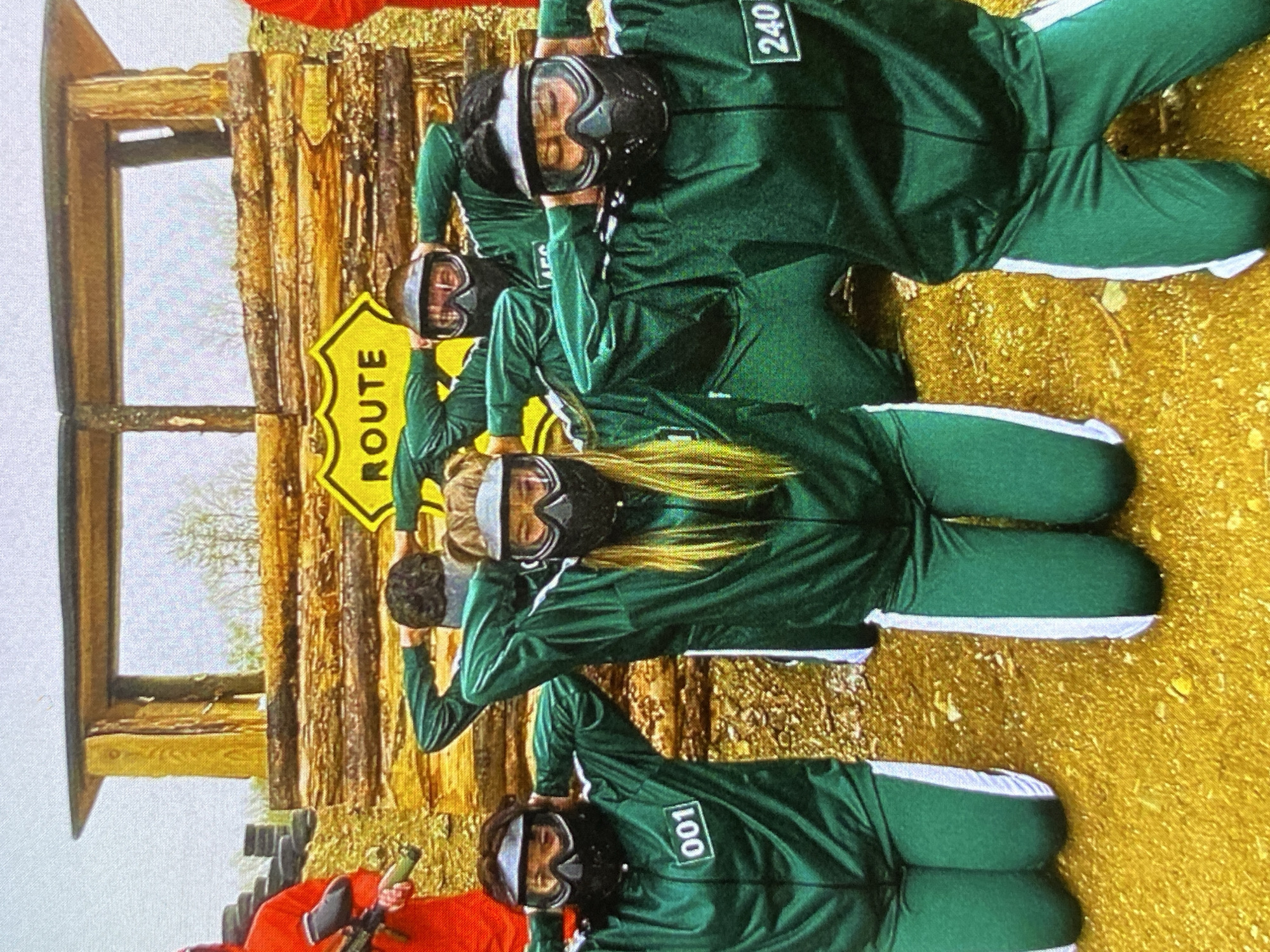 The image captures an outdoor scene in Bankya, Bulgaria, featuring several individuals participating in a themed activity, likely paintball or an adventure game. The primary subjects are four people dressed in dark green tracksuits with white side stripes, resembling uniforms from the "Squid Game" series. Each person wears a black paintball mask or goggles, obscuring their faces. Visible numbers on the tracksuits include "001" and "240," and a partial "AF..." on another. They are seen in various poses, some with hands on their heads, suggesting a game stance or readiness for a photograph.
In the background, a rustic wooden structure built from logs forms an archway or a part of an obstacle course. A yellow, diamond-shaped sign with the word "ROUTE" written vertically in black letters is prominently displayed near this structure. The ground is covered with a yellowish, textured material, possibly dry leaves, straw, or artificial turf, indicating an outdoor setting. In the bottom left, a person wearing a red uniform, also characteristic of "Squid Game," holds a dark-colored paintball marker. The activity appears to be a recreational event or game inspired by the popular show. The lighting suggests it is daytime, possibly under overcast conditions.