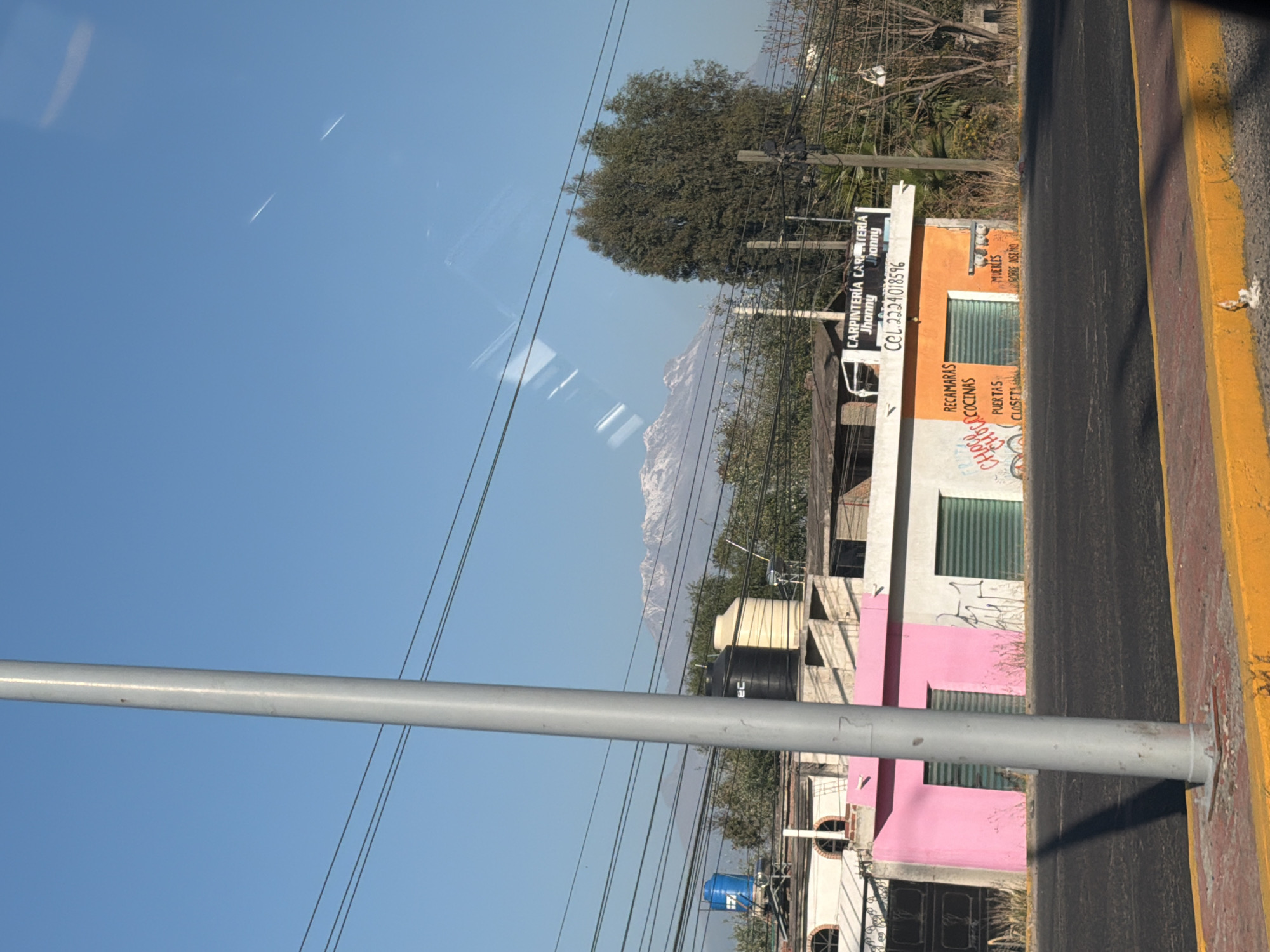 The image depicts a daytime street scene in Santa María Zacatepec, Mexico, with a prominent snow-capped mountain visible in the distance under a clear blue sky. A tall, light gray utility pole stands in the center-left foreground, partially obscuring the view. Behind this pole, a dark asphalt road occupies the mid-ground, bordered by a yellow curb and a reddish-brown sidewalk.
Across the street, two single-story buildings are visible. The building on the left features a pink lower facade with a white upper section and two closed green roll-down security shutters. Graffiti is present on the pink surface. Above this building, multiple water tanks, including one blue, one black, and one cream-colored, are visible on rooftops. Another white building with arched window openings is partially visible behind and to the left of the pink structure.
The building to the right has an orange lower facade with a white upper section, also featuring two closed green roll-down security shutters. Text painted on the orange section reads "RECAMARAS COCINAS PUERTAS CLOSET" and "MUEBLES SOBRE DISEÑO." Above this building, a white sign with black lettering reads "CARPINTERÍA JHONNY" and "CEL. 2224018596." Trees and general vegetation line the area behind the buildings. Multiple dark electrical or communication wires stretch horizontally across the frame at various heights, crossing behind the foreground pole and another utility pole to the right. The distant background reveals a large, snow-covered mountain range partially obscured by atmospheric haze. Reflections are visible on the upper right portion of the image, suggesting the photograph was taken through glass. No individuals are present in the scene.