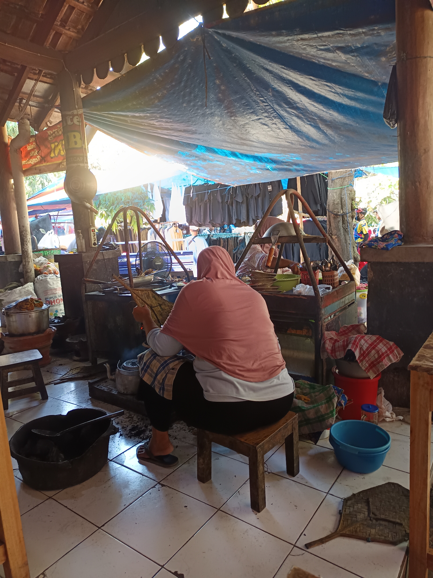 The image depicts a scene at an outdoor food stall in Geneng, Indonesia. A woman, seen from the back, is seated on a wooden stool and appears to be preparing food. She wears a pink headscarf and a light-colored top with dark pants. In front of her is a cooking setup with what looks like a metal stove and various cooking utensils. There are also bags of ingredients, pots, and other food-related items around the stall. Behind her, clothes are hanging on a rack, suggesting this might be a marketplace or a combined food and retail stall. The overall environment is rustic, with a thatched roof overhead partially covered by a blue tarp. The lighting suggests it is daytime, with sunlight filtering through the structure. Other people are visible in the background, possibly customers or vendors, with some wearing helmets, indicating they may have arrived by motorcycle.