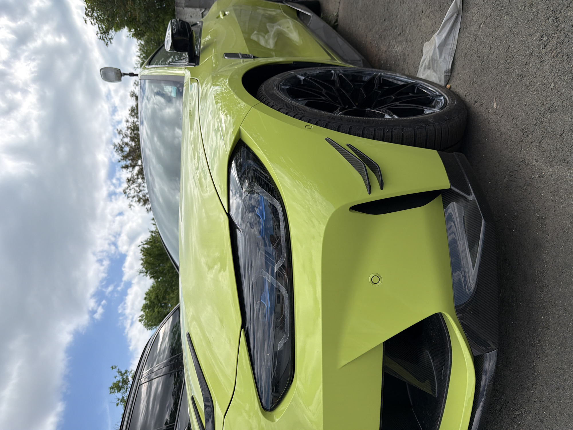 A close-up, vertically oriented shot depicts the front of a bright yellow modern car, likely a BMW M4. The focus is on the headlight, which is black and angular in design. Adjacent to the headlight are two small, dark-colored carbon fiber accents. The car's tire, also black, is visible in the upper right corner. The car is parked on a gray asphalt surface. In the background, a mix of green foliage and a cloudy sky provide context. The location is likely Burgas, Bulgaria.