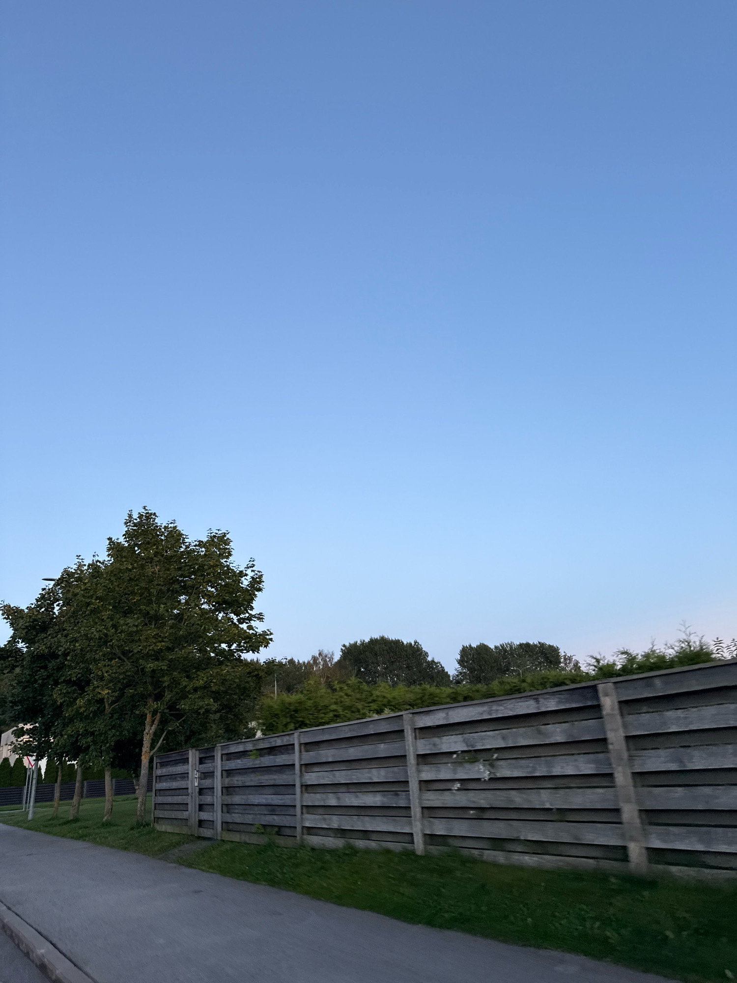 In the city of Alliku, Estonia, an outdoor scene captures a clear sky with a pale blue gradient. The horizon is marked by a dense line of trees and vegetation. In the foreground, a long, grey wooden fence with concrete posts runs horizontally across the image, separating a paved pathway on the left from a strip of green grass on the right. Several trees with dark green foliage stand to the left of the fence, their trunks visible. The overall impression is one of a peaceful, suburban or semi-rural environment, likely in the late afternoon or early evening given the soft lighting. There are no people or animals visible in the frame.
