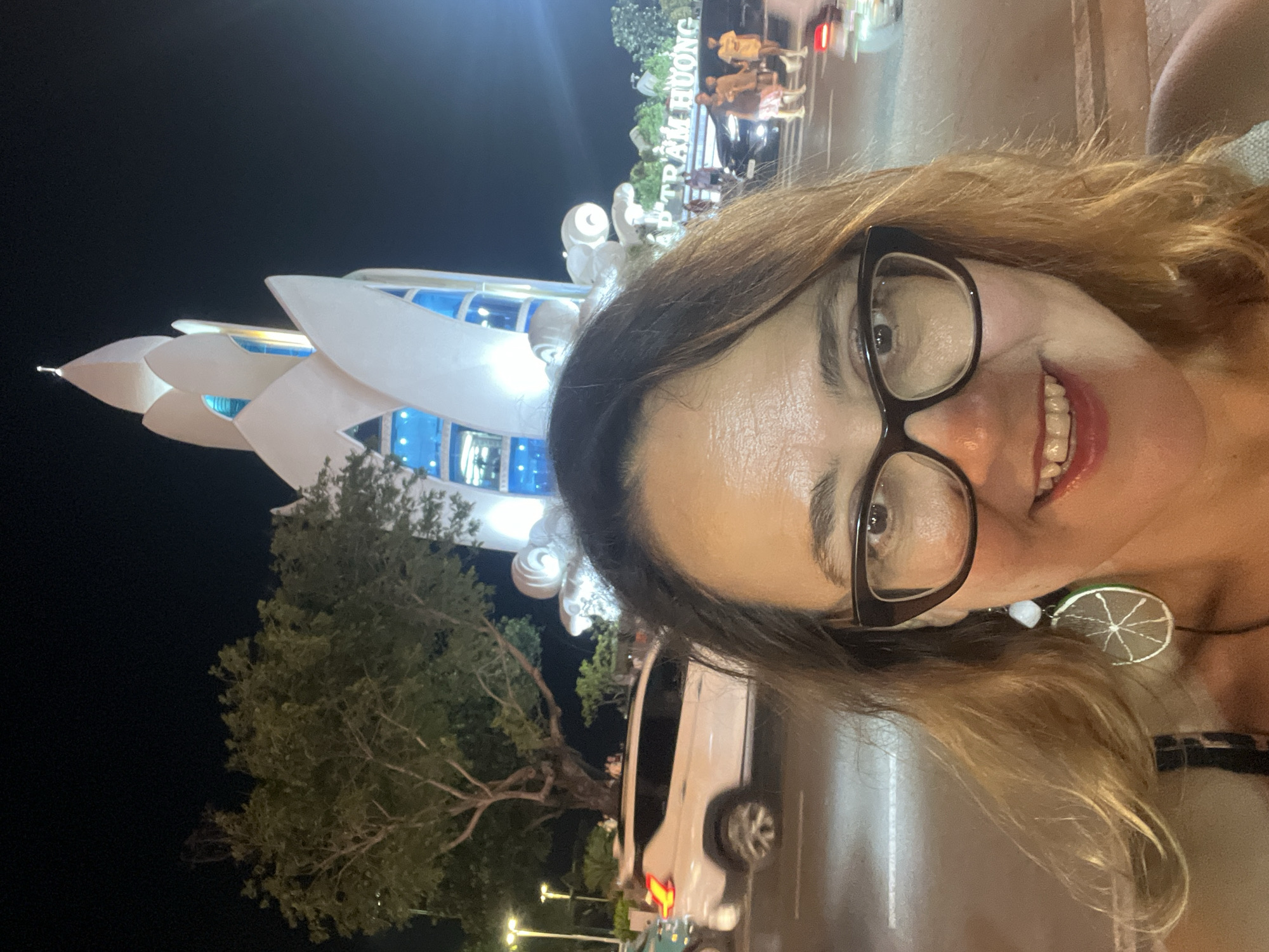The image captures a selfie taken at night in Nha Trang Ward, Vietnam, in front of the Tran Huong tower, a prominent landmark. The central subject is a woman with auburn hair, wearing black-rimmed glasses and a lime-slice-shaped earring. She is smiling directly at the camera. Behind her, the white, lotus-shaped Tran Huong tower is illuminated, with blue lights visible through its windows. To the left of the tower, a large tree with green foliage is visible, and streetlights cast a glow on the scene. In the middle ground, a white SUV is driving on a road, with other cars and pedestrians blurred in the background. A sign with Vietnamese text, "AP TRAM HUONG", is also visible. The overall scene depicts a vibrant nighttime atmosphere in a city setting.
