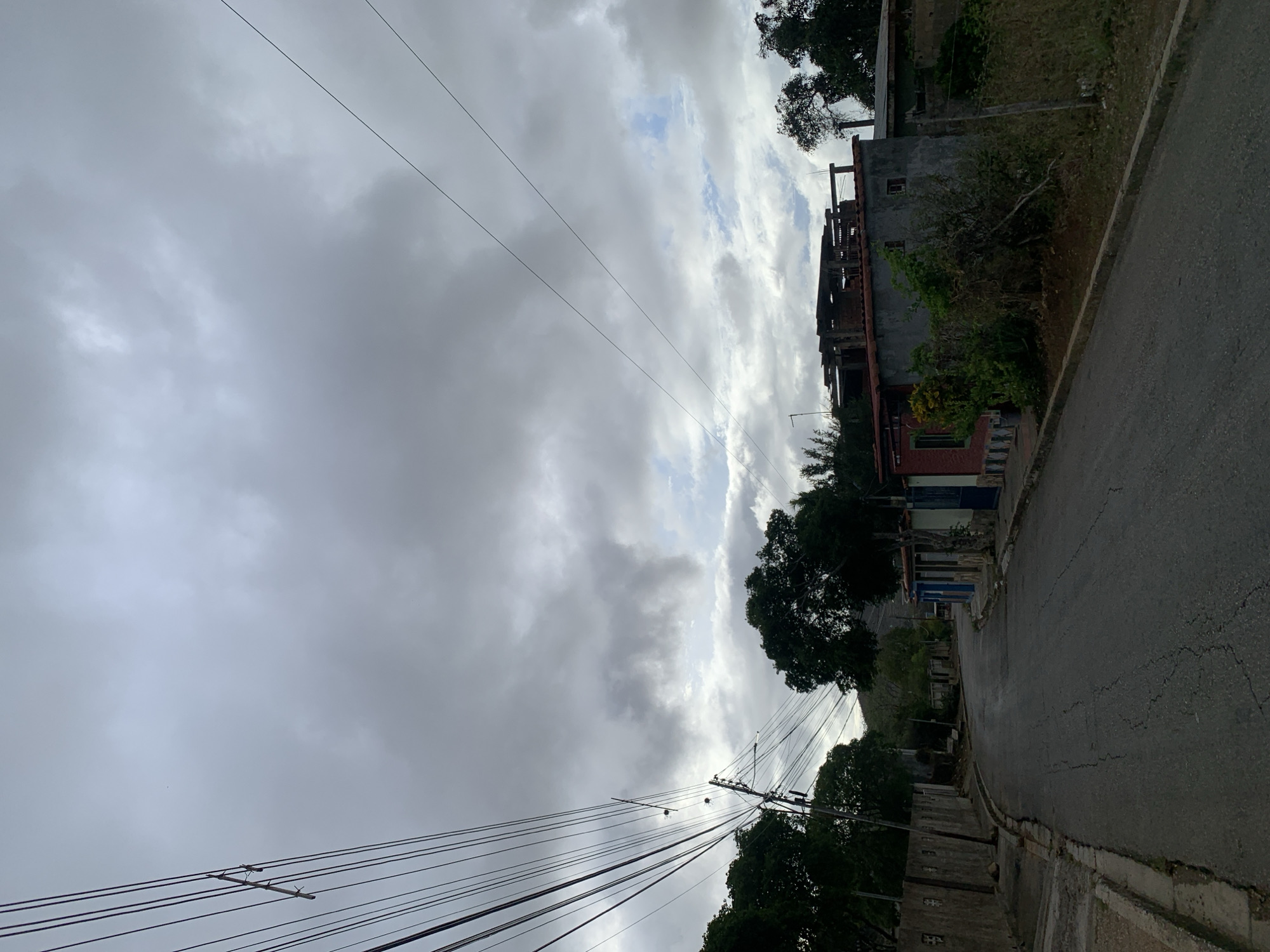 The image captures a street scene in La Tagua, Venezuela, under a heavily overcast sky. The view looks down a curving, asphalt road that appears to descend slightly, flanked by concrete curbs. The road shows some cracks and wear.
The sky dominates the upper portion of the frame, filled with dense, grey clouds, though patches of brighter sky and more textured, lighter clouds are visible, suggesting the sun might be trying to break through or is behind the cloud cover. Numerous overhead power lines crisscross the upper and middle sections of the image, strung between unseen poles, adding to the urban infrastructure feel.
Along the right side of the road, several buildings are visible. Some appear to be residential structures, while others show signs of incomplete construction, with exposed concrete and rebar. One building has a partially red-colored facade and a dark roof. Lush green trees and various bushes are interspersed along the roadside and around the buildings, adding natural elements to the scene. In the distance, vague outlines of hills or mountains can be discerned through the cloudy haze.
No people, vehicles, or discernible activities are taking place, leaving the scene tranquil and focused on the street, buildings, and sky. No visible text is present. The general atmosphere is one of a quiet, developing residential area on a cloudy day.