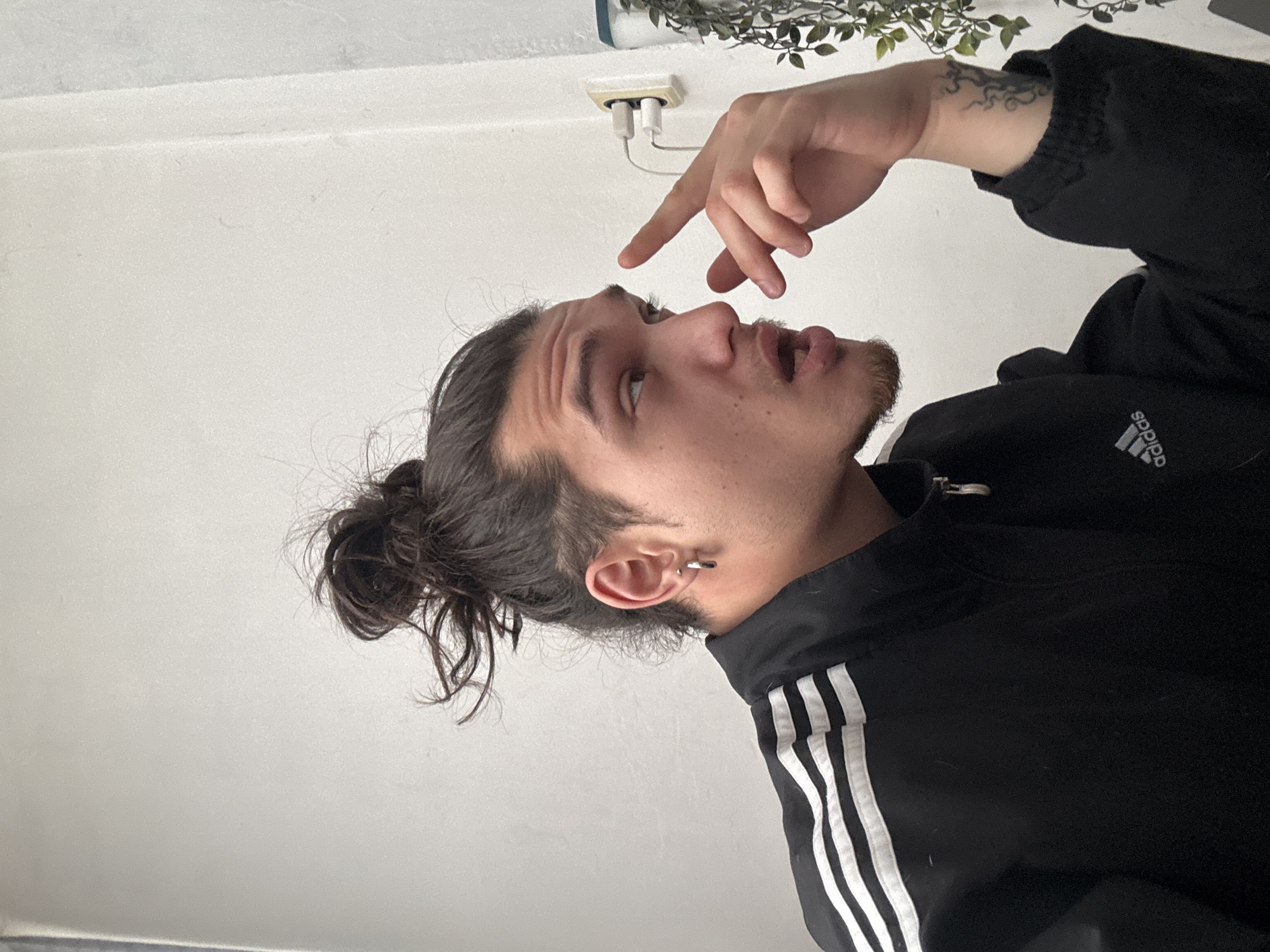 A medium shot depicts a man in a black Adidas jacket. He has dark hair tied in a bun and a small hoop earring in his left ear. He is looking up and to the left, pointing at his nose with the index finger of his right hand. The man has a mustache and a light beard. Above and to the left of him are a wall, a leafy potted plant, and a white electrical outlet with two phone chargers plugged into it. The location is unidentified; there is no visible evidence to determine this image was taken in Sofia, Bulgaria.