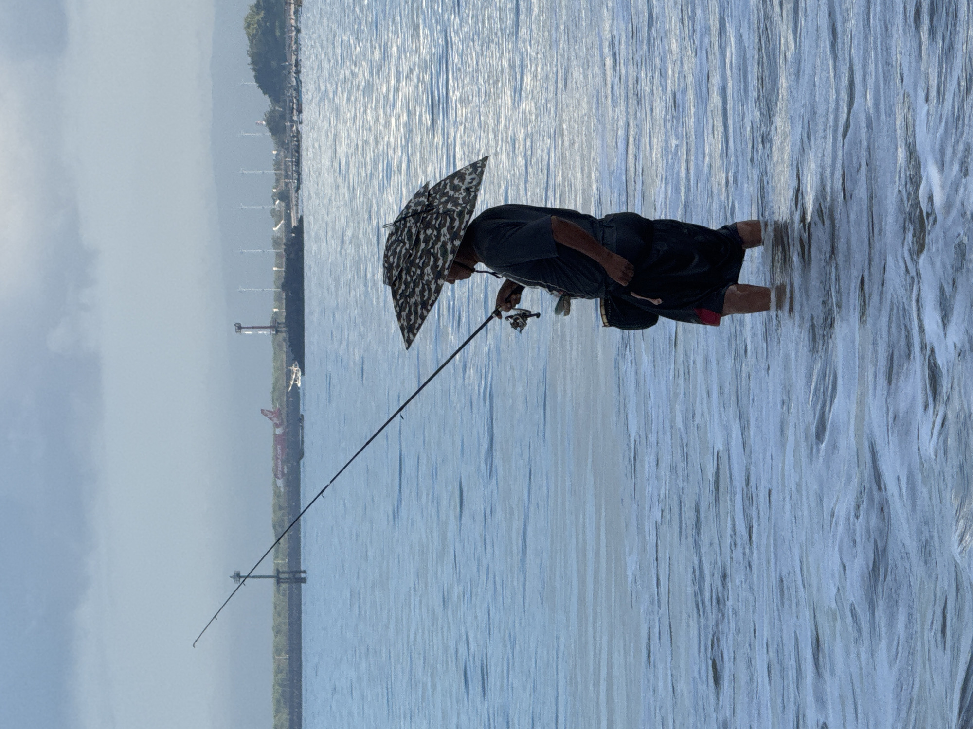 A man is fishing in shallow water along the shoreline of Kuta, Indonesia. He is standing in the water, up to his mid-calf, holding a fishing rod. He is also holding an umbrella over his head. In the background, there is a coastline with buildings and structures visible. The water is calm and reflective, with subtle wave patterns. The sky is overcast.