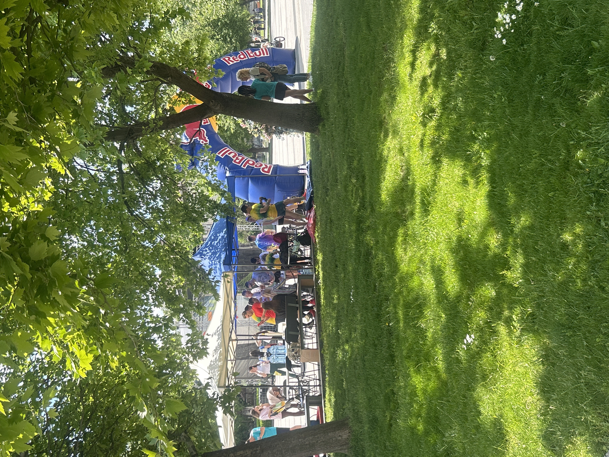 Outdoors on a sunny day in Pleven, Bulgaria, a partially shaded grassy area is adjacent to a Red Bull event. A tree provides shade to a portion of the grass and the Red Bull setup. People are near the Red Bull structure, and some appear to be interacting with the event. The setting suggests an outdoor promotional event.