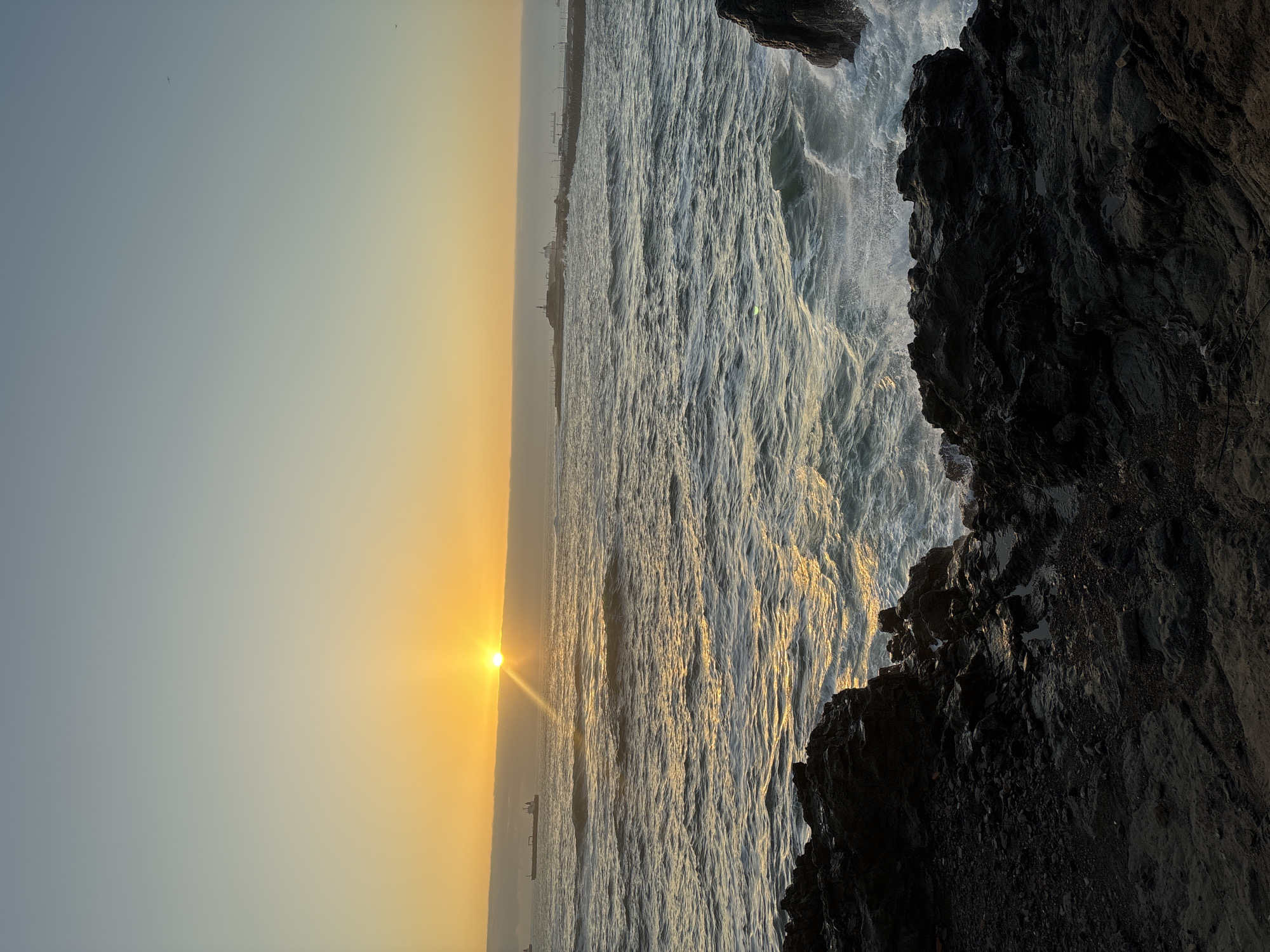 The photograph, taken in Arica, Chile, depicts a coastal scene at sunset. The sun is visible near the horizon, casting a bright light and warm golden hue across the water and sky. The sea is turbulent, with visible waves and foam, suggesting possible rough conditions. Dark, rocky formations are prominently featured on the right side of the frame, indicating a rocky coastline. In the distance, faint outlines of buildings and structures can be seen along the coastline, representing the urban environment of Arica. A lone vessel is visible on the water. The sky is a gradient of pale blue to orange, with no other visible details.