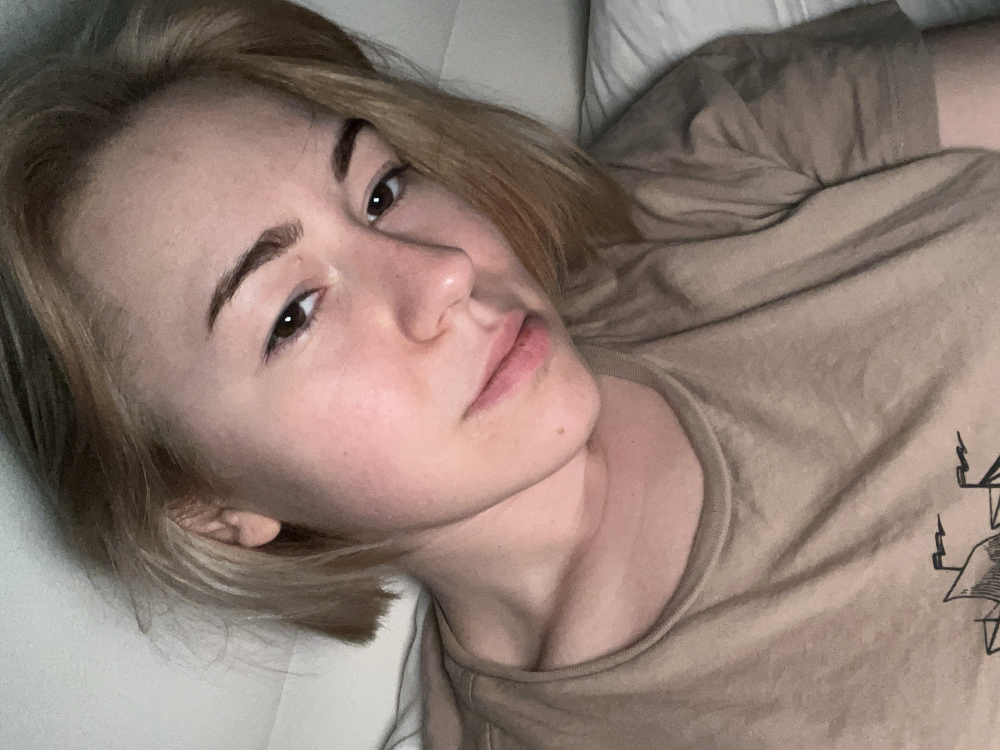 A young woman with short, blonde hair and brown eyes is lying down, likely on a bed, as suggested by the white pillow visible in the background. She is wearing a brown t-shirt with a black graphic design on the right side of her chest. Her skin tone is fair, and she has slightly arched eyebrows. The lighting is soft and somewhat diffused. No location in Boreti, Montenegro is discernible from the image.