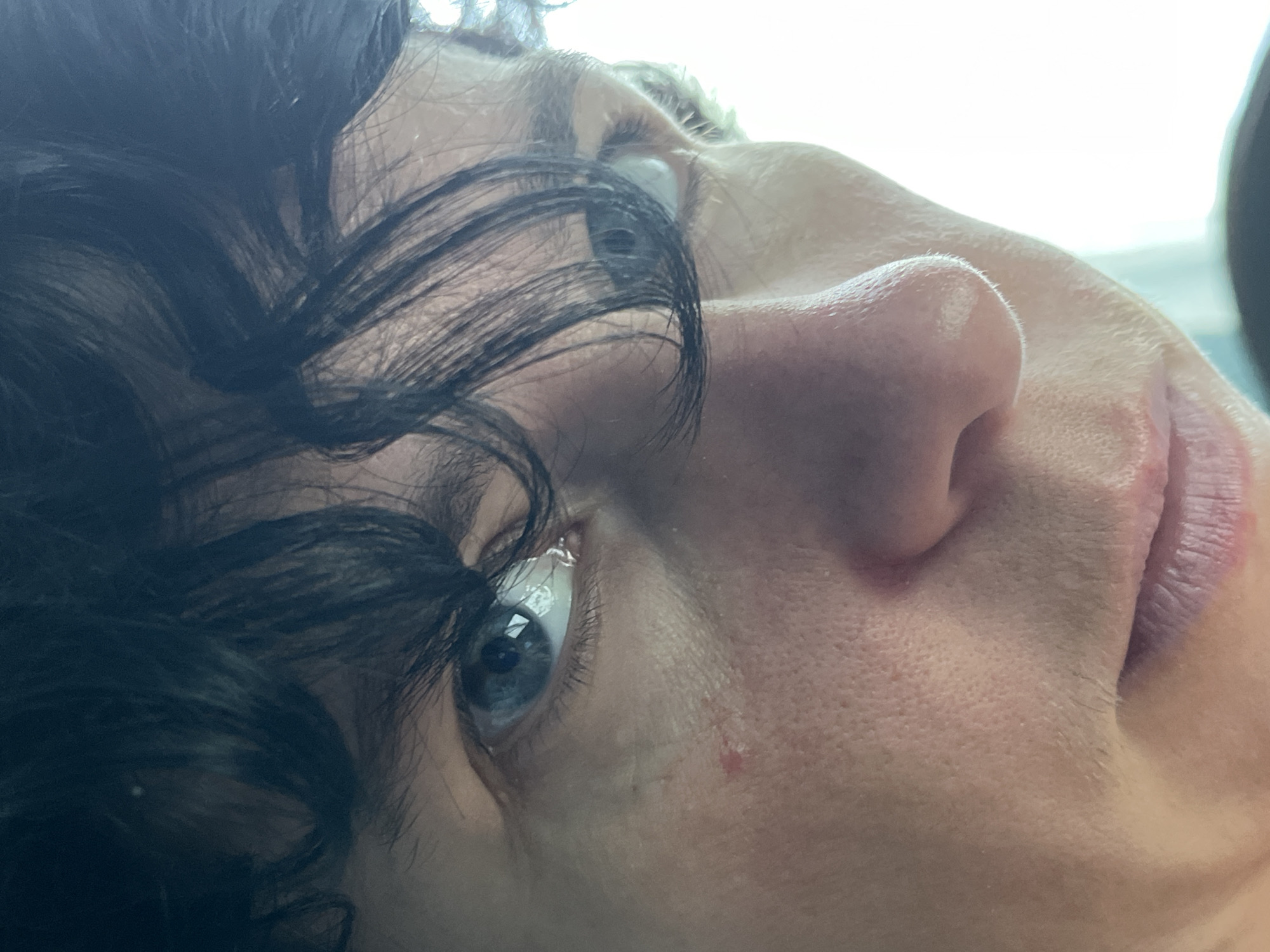 The image is a close-up, low-angle shot of a person's face, focusing on their left eye, nose, and mouth. The person has dark, curly hair that partially obscures their forehead and eyebrows. Their eye is blue with a reflection of a bright light source. The skin has visible pores and a small, reddish mark on the cheekbone. The lighting suggests the subject is near a window or a bright light source, with the background being overexposed and featureless. There is no identifiable information about the location within ღრმაღელის თემი, Georgia, as the image focuses solely on the facial features of an individual without any contextual environmental details.