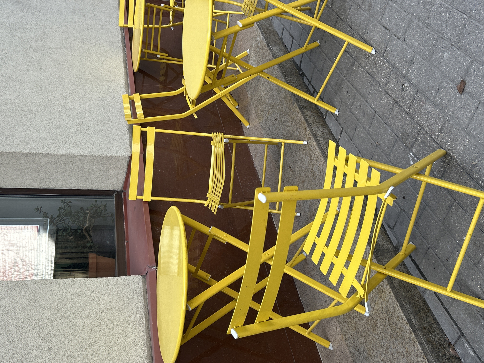 The image displays an outdoor seating area featuring numerous bright yellow metal chairs and matching round tables, arranged alongside a building. The furniture, likely designed to be foldable, appears to be set up on a sidewalk or patio.
Several yellow chairs are prominently visible, some standing upright, while others are positioned at various angles, suggesting they might be partially folded or casually arranged. The chairs have slender metal frames and slatted backrests. Scattered among the chairs are several yellow round tables. White plastic caps are visible on the feet of many chairs, protecting the ground surface.
The setting is an urban or commercial outdoor space. Adjacent to the seating area is a building with a textured, light gray facade at its lower level. Above this, a large window with a dark frame reflects blurred imagery, possibly showing indoor plants or structures across the street. The ground beneath the furniture consists of two distinct surfaces: a smooth, dark reddish-brown area immediately next to the building, and a section of gray, rectangular brick-patterned pavement extending further out.
The scene is brightly lit, indicative of daytime, with soft, even lighting and no harsh shadows. No people are present, suggesting the area is unoccupied or closed for the moment. There is no visible text in the image. The vibrant yellow color of the furniture creates a cheerful and inviting atmosphere.