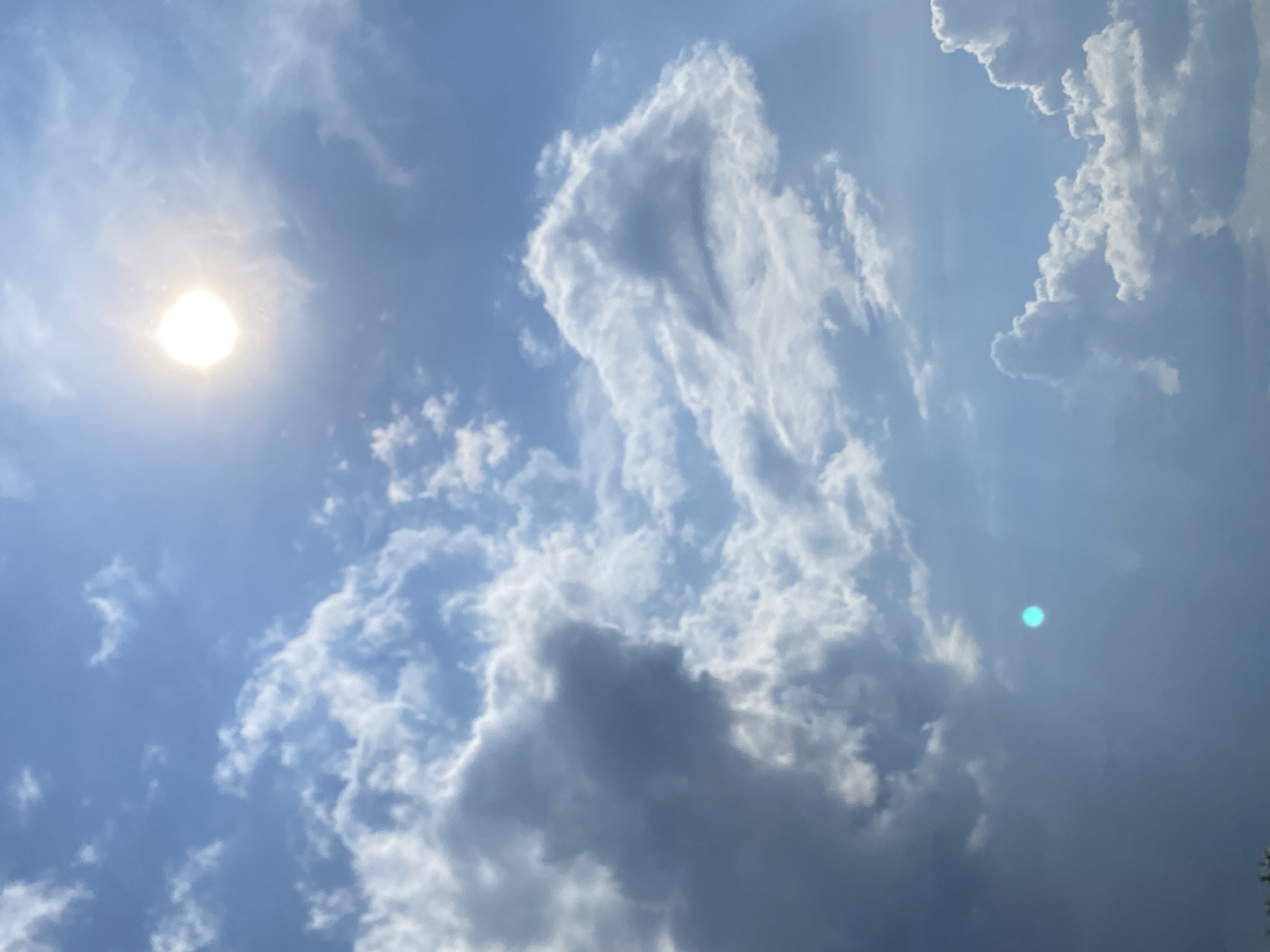 The image depicts a bright, sunny sky with scattered clouds. The sun is visible as a bright orb in the upper right quadrant of the image, with a slight lens flare effect appearing as a turquoise circular light in the lower left quadrant. The clouds are a mix of fluffy white cumulus clouds and darker, more diffused grey clouds, suggesting a dynamic weather pattern. The overall scene is one of a clear day with some cloud cover. No specific landmarks or elements identifiable as being in Chaikovsky, Russia, are present in the image. The image is a close-up of the sky and clouds, with no ground features or human presence visible.