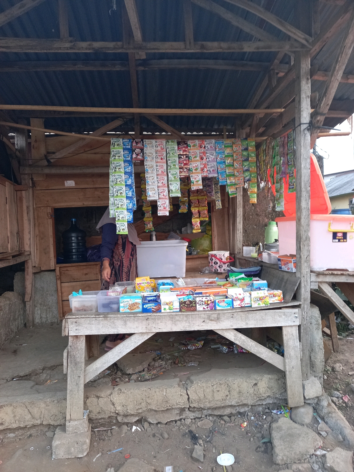 This is a forensic, matter-of-fact description of a small roadside stall selling various packaged snacks and drinks, located in the city of Wonosari, Indonesia. The stall is constructed from rough wooden planks and corrugated metal, with a simple wooden table displaying the merchandise.
**Objects:**
* **Merchandise:** A wide array of small, colorful packaged snacks and drinks are hanging from strings above the table and displayed on top of it. These include items with text in what appears to be Indonesian or Malay. Prominent brands or types of items visible include those labeled "OPPA," "Frizzy," "Oreo," and "Bello Waffle."
* **Table and Shelving:** A rustic wooden table, supported by wooden posts and concrete blocks, holds some of the merchandise in clear plastic containers and boxes. A wooden shelf structure is behind the table.
* **Water Dispenser:** A large black water jug is visible inside the stall's wooden structure.
* **Cooler/Storage Boxes:** A large white plastic storage box and a smaller pink one are present, one of which has text printed on it.
* **Person:** A person wearing a headscarf and traditional patterned clothing is partially visible behind the table, attending to the stall. Their hands are visible, suggesting they are interacting with the items.
* **Environment:** The stall is situated on uneven, dirt and concrete ground. Debris and scattered litter are visible on the ground, suggesting a less maintained area. In the background, there are other simple structures, one with a corrugated metal roof and another with a bright orange roof.
**Actions and Interactions:**
The person at the stall appears to be an attendant, likely organizing or selling the goods. The display of snacks suggests a point of sale.
**Environment Details:**
The construction materials, the type of merchandise, and the overall presentation point to a typical small, informal retail business found in a developing urban or peri-urban area of Indonesia. The presence of debris indicates a busy, possibly high-traffic area where litter accumulates. The background elements suggest a neighborhood with a mix of residential and commercial structures.