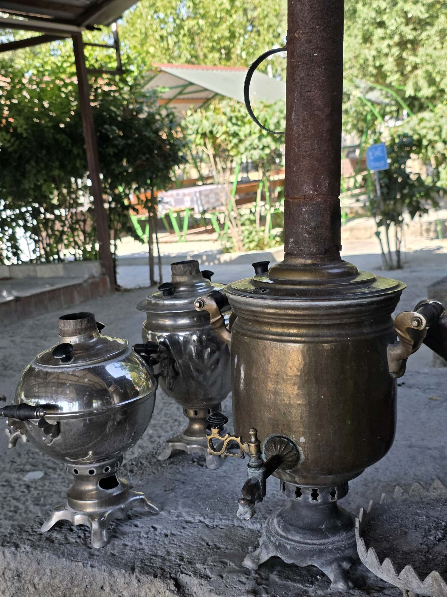 Three samovars, traditional Russian tea urns, are displayed outdoors on a paved surface. The samovar in the foreground on the right is made of brass and features a prominent tap. To its left, a smaller, polished metal samovar sits on a pedestal with intricate legs. In the background, another polished metal samovar is visible. The setting appears to be a park or garden area, with lush green trees and foliage surrounding the samovars. In the distance, tables and chairs are set up under a canopy, suggesting a seating area. The overall atmosphere is serene and offers a glimpse into a cultural display, possibly in the city of Qobustan, Azerbaijan, known for its historical significance and natural beauty.