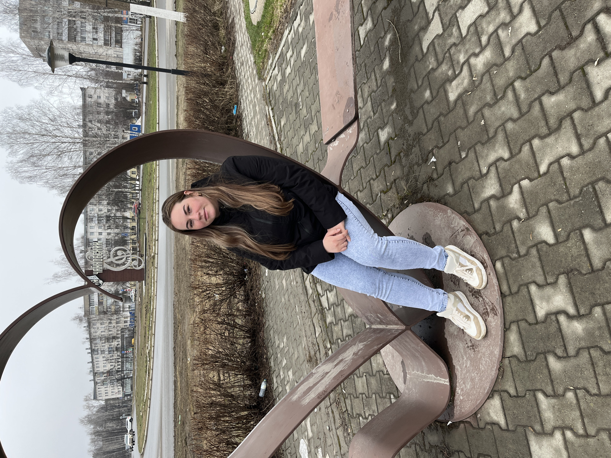 A young woman is sitting on a metal sculpture that appears to be a public art installation in the city of Chaikovsky, Russia. The sculpture has a rusty brown color and a curved, open design. She is wearing a black jacket, light blue jeans, and white sneakers with tan accents. She is sitting with her legs extended and her hands in her lap. The sculpture is located on a paved area with a brick pattern. In the background, there is a road, a grassy area with bare bushes, and several buildings. The sky is overcast. There is also a tall lamp post visible, as well as another similar sculpture with musical notes nearby.