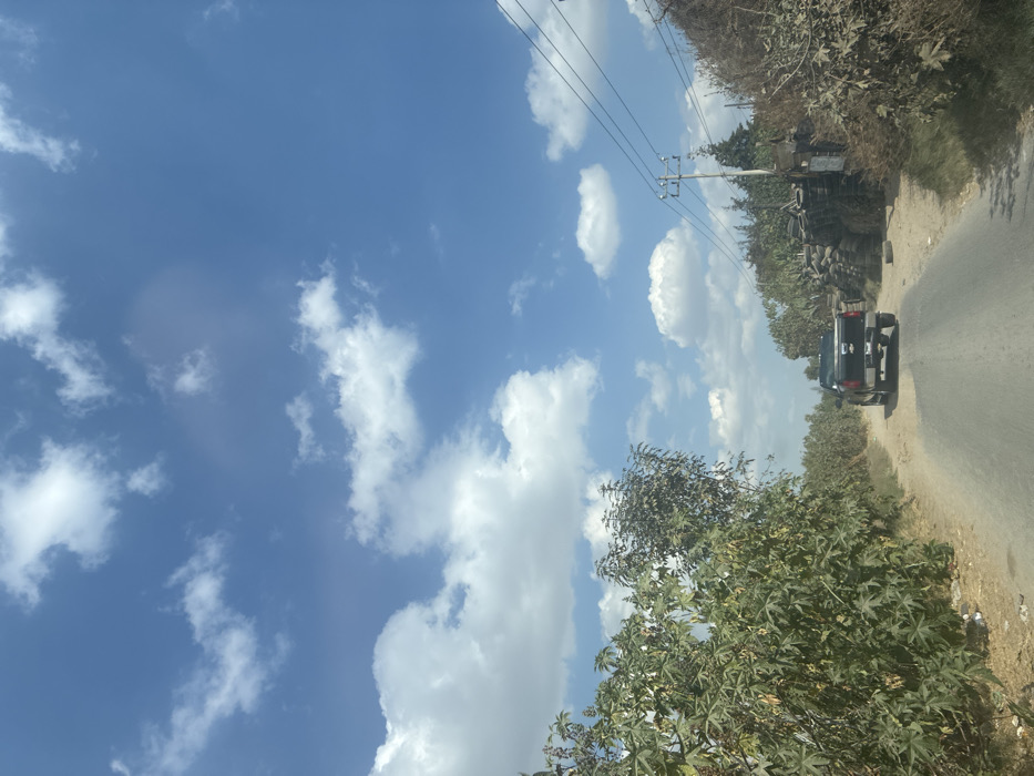 A road cuts through a landscape under a bright blue sky dotted with fluffy white clouds. A dark-colored pickup truck drives away on the asphalt. Vegetation, including trees, lines both sides of the road. Power lines run overhead, supported by poles. Various objects, possibly tires, are piled up near one of the poles. The scene suggests a rural or semi-rural setting.