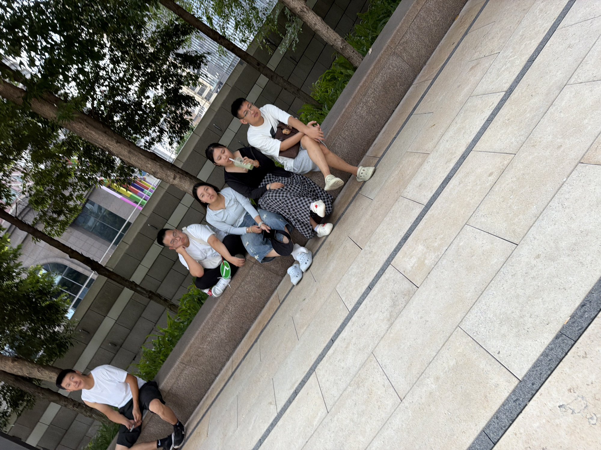The image depicts a group of five individuals, three males and two females, seated on a stone ledge outdoors. The setting appears to be a public space in Yuzhong District, China, with trees and modern buildings visible in the background.
On the far left, a male wearing a white sleeveless shirt and black shorts is seated. Next to him, a male in a white t-shirt and dark pants is also seated. To his right, a female in a light blue top and jeans is seated. Adjacent to her, another female in a black top and a plaid skirt is seated, holding a green beverage. The final individual on the right is a male wearing a white t-shirt, grey shorts, and glasses. He is holding a phone. All individuals appear to be relaxed.
The ground in front of the ledge is paved with light-colored rectangular tiles arranged in a diagonal pattern, with dark gray strips separating the rows. The ledge itself is made of a reddish-brown stone. The overall impression is that of a casual gathering in an urban environment.