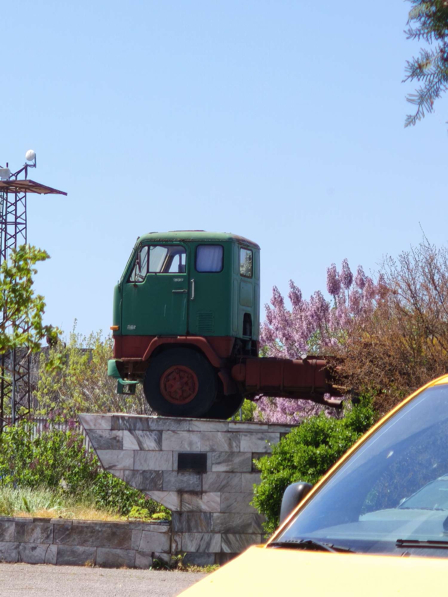 An old, green and rust-colored truck cab and chassis are prominently displayed as a monument on a multi-tiered stone pedestal under a clear, bright blue sky in Burgas, Bulgaria. The truck, a cab-over-engine model, features a green cab with a rusty reddish-brown lower chassis and wheel. Faint text, "U4850", is visible on the driver's side door, and "858" appears below the windshield. A small white curtain hangs inside the cab's window.
The pedestal is constructed from light-colored, rectangular stone blocks, possibly marble or granite, arranged in an angular, stepped design. To the left of the monument, a metal lattice structure, possibly a communication tower, is partially visible, surrounded by green foliage. To the right, vibrant bushes with pink and purple blossoms, suggesting spring, are alongside trees with bare or brown branches. In the bottom right foreground, the yellow hood and windshield of a vehicle are partially visible. The scene is brightly illuminated by daylight, indicating clear weather.