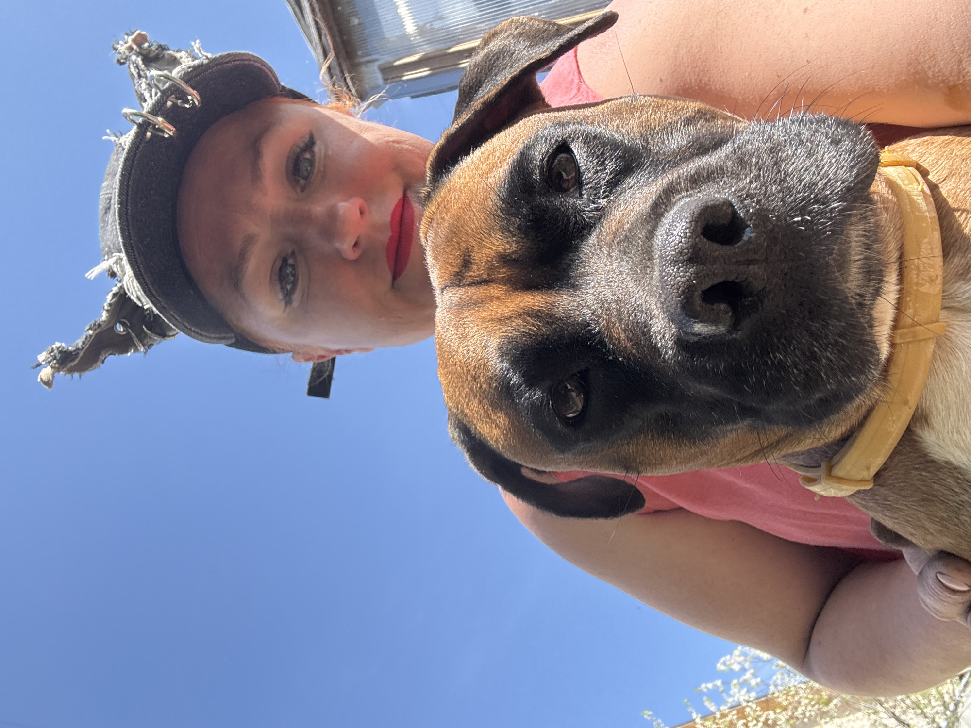 A woman in a dark, fringed hat and red lipstick holds a brown boxer dog. The woman has fair skin and is looking at the camera. The dog has a yellow collar. The background is a bright blue sky. The location is Chaikovsky, Russia.