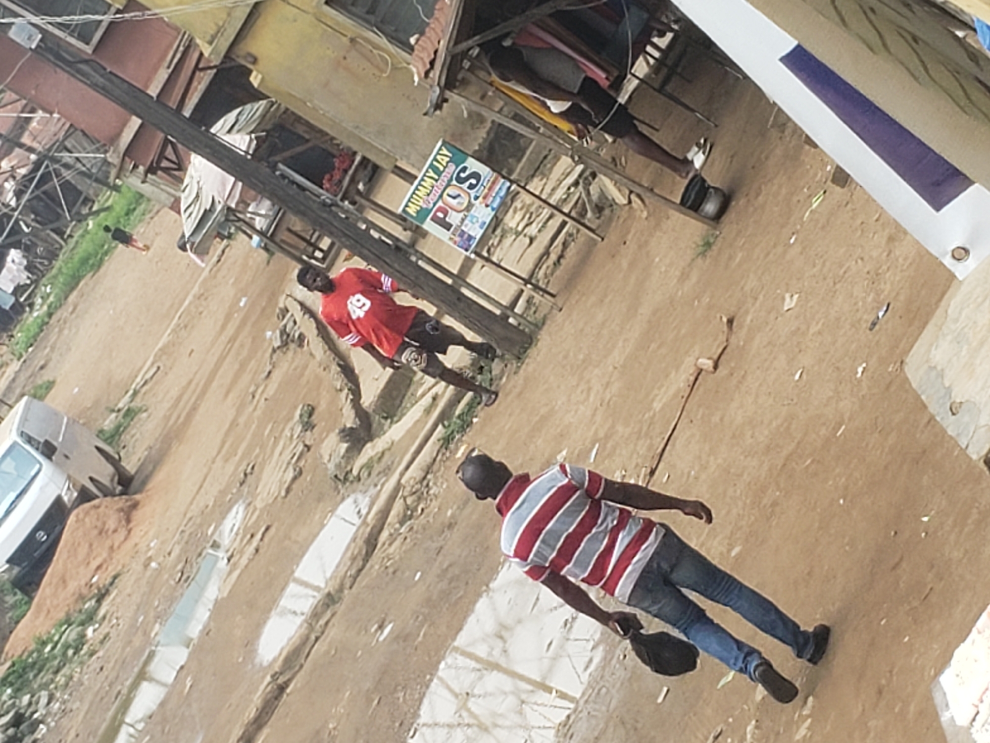 The image captures a daytime street scene in Alalubosa, Nigeria, showing a dusty, unpaved road with scattered debris and several reflective wet patches or puddles.
In the foreground, a man with a shaved head is seen from behind, walking away from the camera. He wears a red and white striped polo shirt, blue jeans, and dark shoes, and carries a black bag in his left hand. Further into the midground, another man stands, partially facing away, dressed in a red t-shirt with the number "49" on the back and dark shorts.
To the left, a white van or minibus is partially visible, positioned near a mound of reddish-brown dirt. The background features several informal structures, likely small shops or stalls, constructed from wood and simple materials, with one building having a yellow facade. Inside one of these structures, a person's legs and feet are visible near a black pot on the ground.
A utility or electrical pole cuts diagonally across the frame. Attached to this pole is a green and white sign that clearly reads "MUMMY JAY" at the top. Below this, "POS" (likely Point of Sale) is prominently displayed in large letters, with smaller text that appears to be "Wallets" and various logos for mobile money or financial services underneath. The scene depicts a bustling, semi-urban environment with pedestrian activity and small-scale commerce.