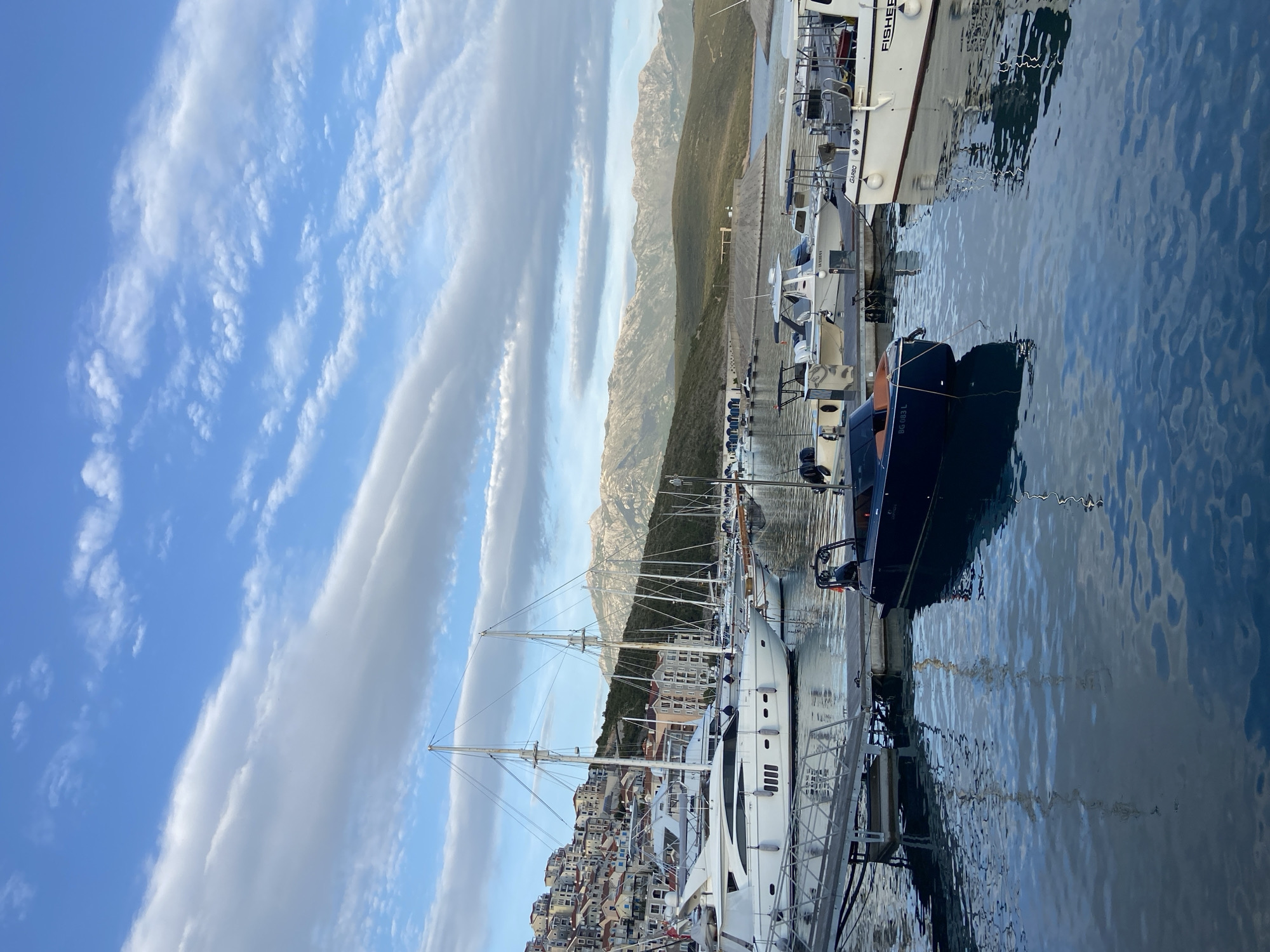 The image captures a picturesque marina scene in Radovići, Montenegro, under a clear sky. In the foreground and middle ground, numerous boats of various sizes are docked along piers. A large, elegant white yacht with multiple masts is prominent on the left side of the marina. To the right, several smaller white motorboats and a dark blue boat, bearing the text "BC 083 L," are moored. Another larger white boat in the upper right has "FISHER" and "G480" visible on its hull. The calm, blue water of the marina reflects the boats and the sky, showing gentle ripples.
Beyond the marina, a densely packed town or village with light-colored buildings cascades down a hillside. These structures ascend towards the middle ground, where rugged, rocky mountains form a dramatic backdrop, stretching horizontally across the scene. Some mountain slopes appear barren, while others show sparse vegetation.
The sky overhead is a vibrant blue, adorned with distinctive white clouds arranged in long, parallel, wispy bands, giving the impression of a patterned sky. The lighting in the scene is soft and clear, suggesting either early morning or late afternoon, creating long shadows and contributing to a serene and tranquil atmosphere. No specific human activity is discernable, emphasizing the peaceful setting.