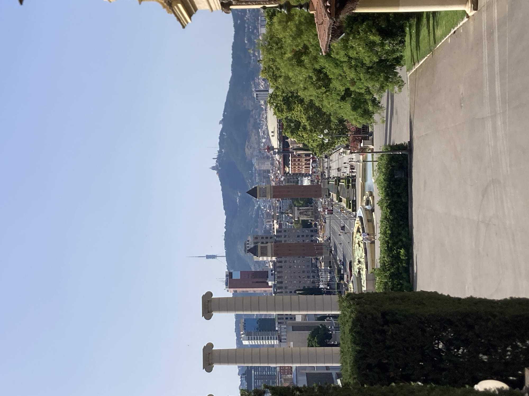The image depicts a panoramic view of Barcelona, Spain, from a vantage point overlooking the Plaça d'Espanya. In the foreground, two imposing Corinthian columns stand tall, framing the scene. Beyond them, a wide promenade stretches towards the city, lined with lush green hedges and a paved pathway. Several people are visible walking along the promenade.
The middle ground features the iconic Venetian Towers, two distinctive brick towers flanking the avenue. A circular fountain with lush greenery and water jets is situated in front of the towers. Cars and buses are observed moving along the busy street.
In the background, the cityscape of Barcelona unfolds with various modern buildings and older structures, including a prominent red-brick building. Further in the distance, a mountain range is visible, topped by several structures, including the Montjuïc Communications Tower. The sky is clear and blue, indicating a sunny day. The overall scene captures a vibrant urban landscape with historical and modern architectural elements under a bright sky.