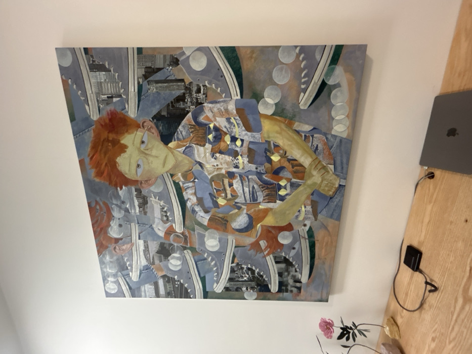 An abstract painting dominates the scene. It depicts a stylized figure with red hair and pale skin, wearing a patterned shirt. The background is a mix of cityscapes, sneakers, and circles. The painting is hung on a white wall, with a wooden desk visible on the right. A laptop and charger sit on the desk, and a small vase with pink flowers is visible below the painting.