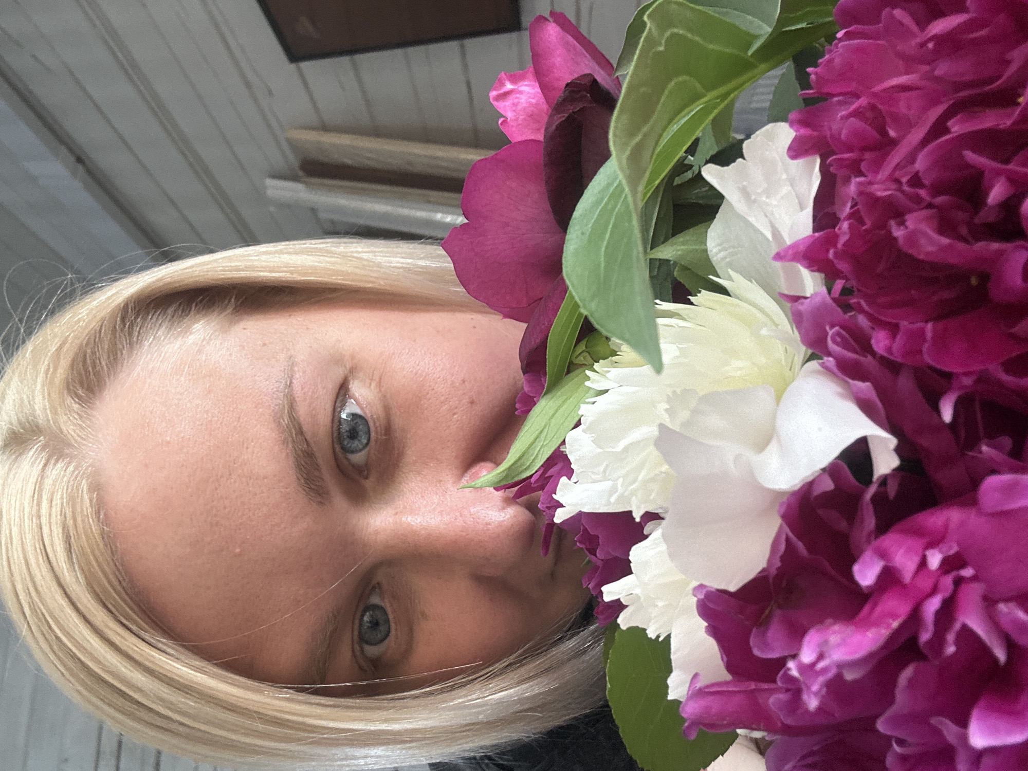The image is a close-up, selfie-style photograph of a woman holding a bouquet of peonies. The woman has blonde hair and blue eyes, and she is peeking over the flowers, looking directly at the camera. The peonies are a mix of deep pink, magenta, and white. The background appears to be a wooden wall, possibly in an outdoor setting. There is no identifiable urban context that would place this image specifically in Chaikovsky, Russia.