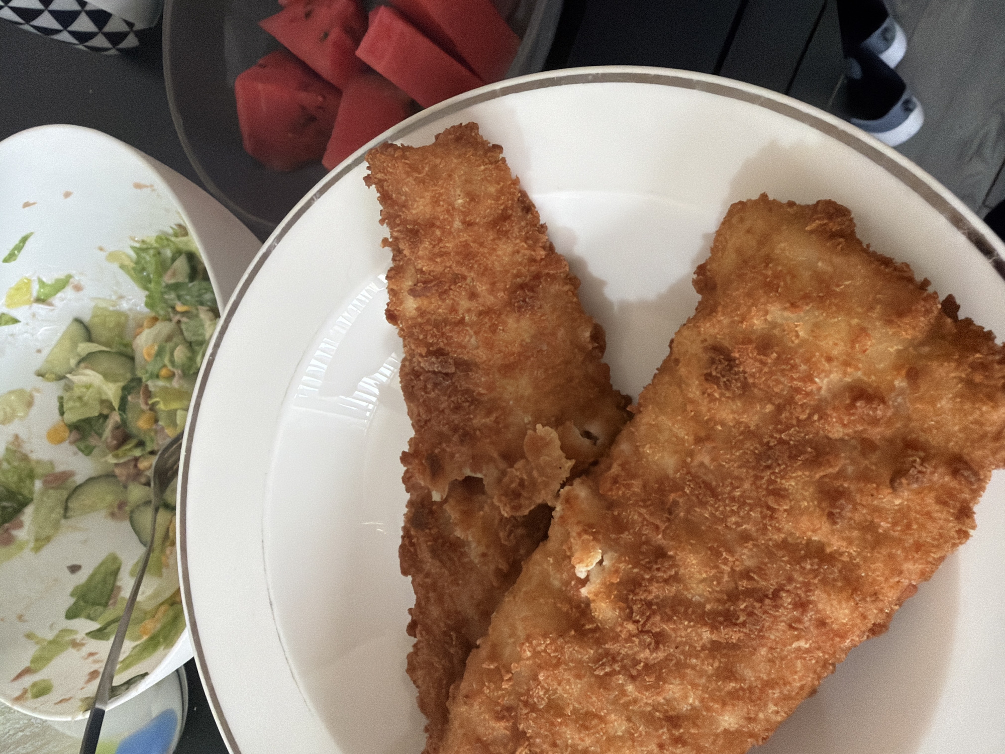 The image shows a meal laid out on a surface, possibly a table. A white plate holds two pieces of golden-brown fried fish. To the left, there's a white bowl containing a salad, and silverware rests inside. Behind the fish, there's a bowl with slices of watermelon. A patterned item and a pair of shoes are visible in the background. The location appears to be Pentre, United Kingdom. There are no people immediately visible, but the arrangement of the food suggests an individual is preparing or has consumed the meal.