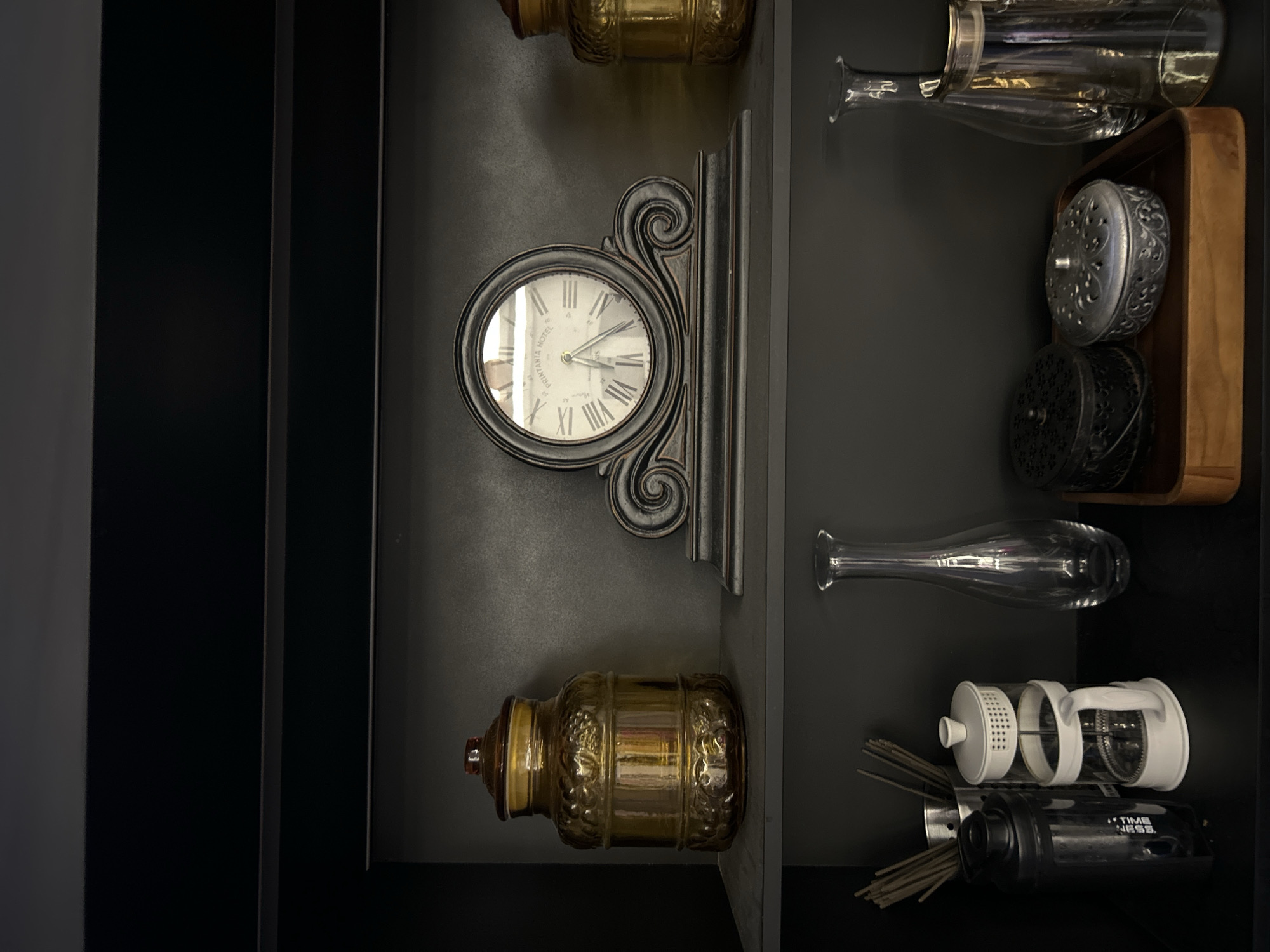 A high-angle shot shows a dark wooden shelf in what appears to be a home in North Kuta, Indonesia, displaying several antique-style items. On the top shelf, a large, ornate amber glass jar with a lid sits to the left of a dark, decorative mantel clock. To the right of the clock is another amber glass jar. The bottom shelf features a black shaker bottle and a white French press coffee maker on the left. To the right of these, a tall, clear glass vase stands next to a wooden tray holding two decorative metal boxes. A clear glass carafe with a silver rim is positioned behind the metal boxes. The overall impression is of a curated collection of decorative objects on a dark shelf.
