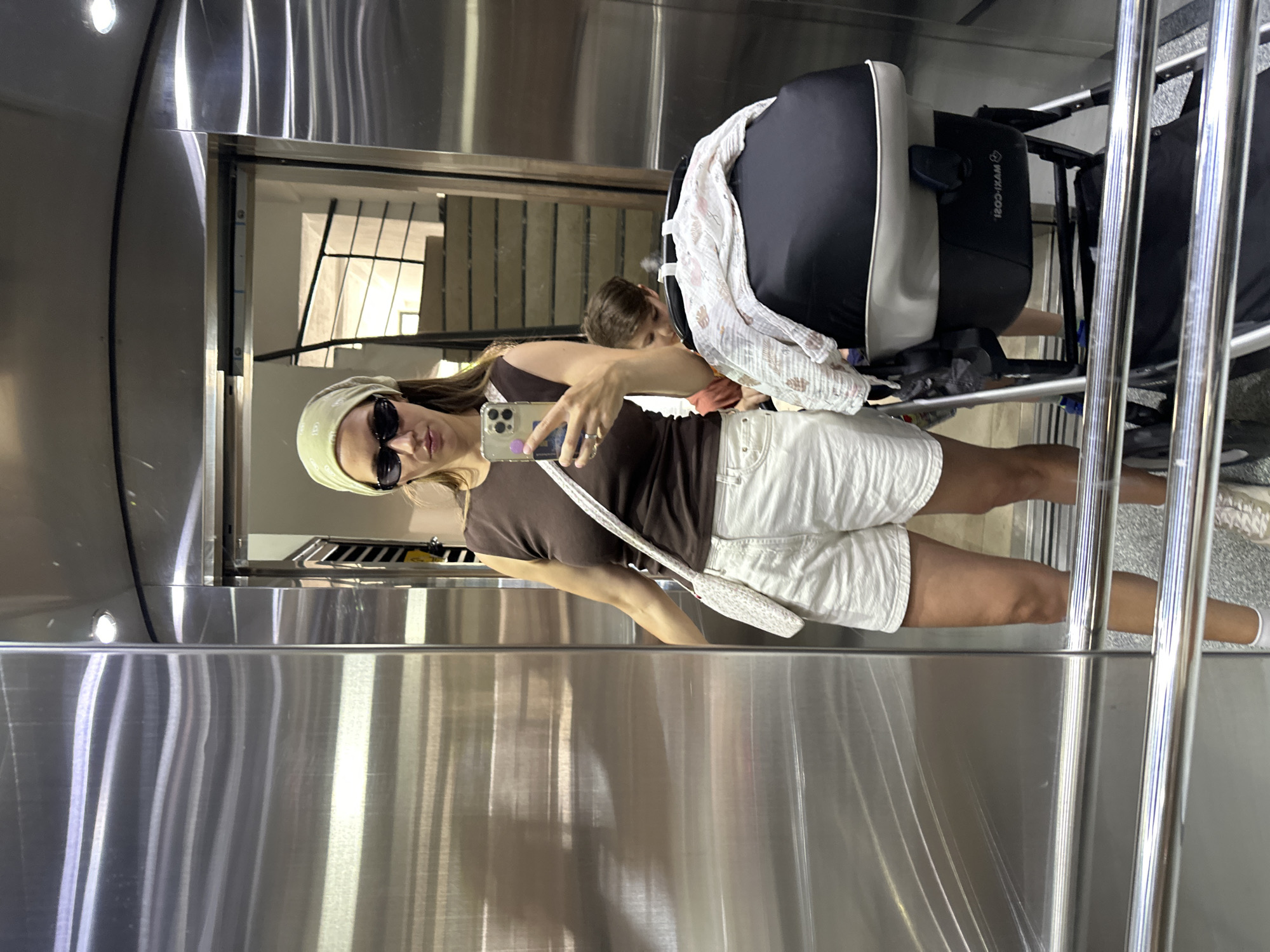 A woman is taking a selfie in a highly reflective elevator, likely using a mirror on one of the walls. She is positioned slightly to the left of the frame, holding her smartphone up to capture the image. She wears a brown t-shirt, white shorts, a light-colored headband, and dark sunglasses, with a white patterned shoulder bag across her body.
Behind her, partially visible, is a child with brown hair, and next to the child is a baby stroller with a black and white padded car seat. The car seat clearly displays the reversed text "MAXI-COSI". The elevator interior appears to be made of polished stainless steel or chrome, creating strong reflections of the woman, the stroller, and the hallway or interior space outside the elevator doors, which feature a patterned design. The overall scene suggests travel or movement within a building. The location context for this image is Fethiye, Turkey.