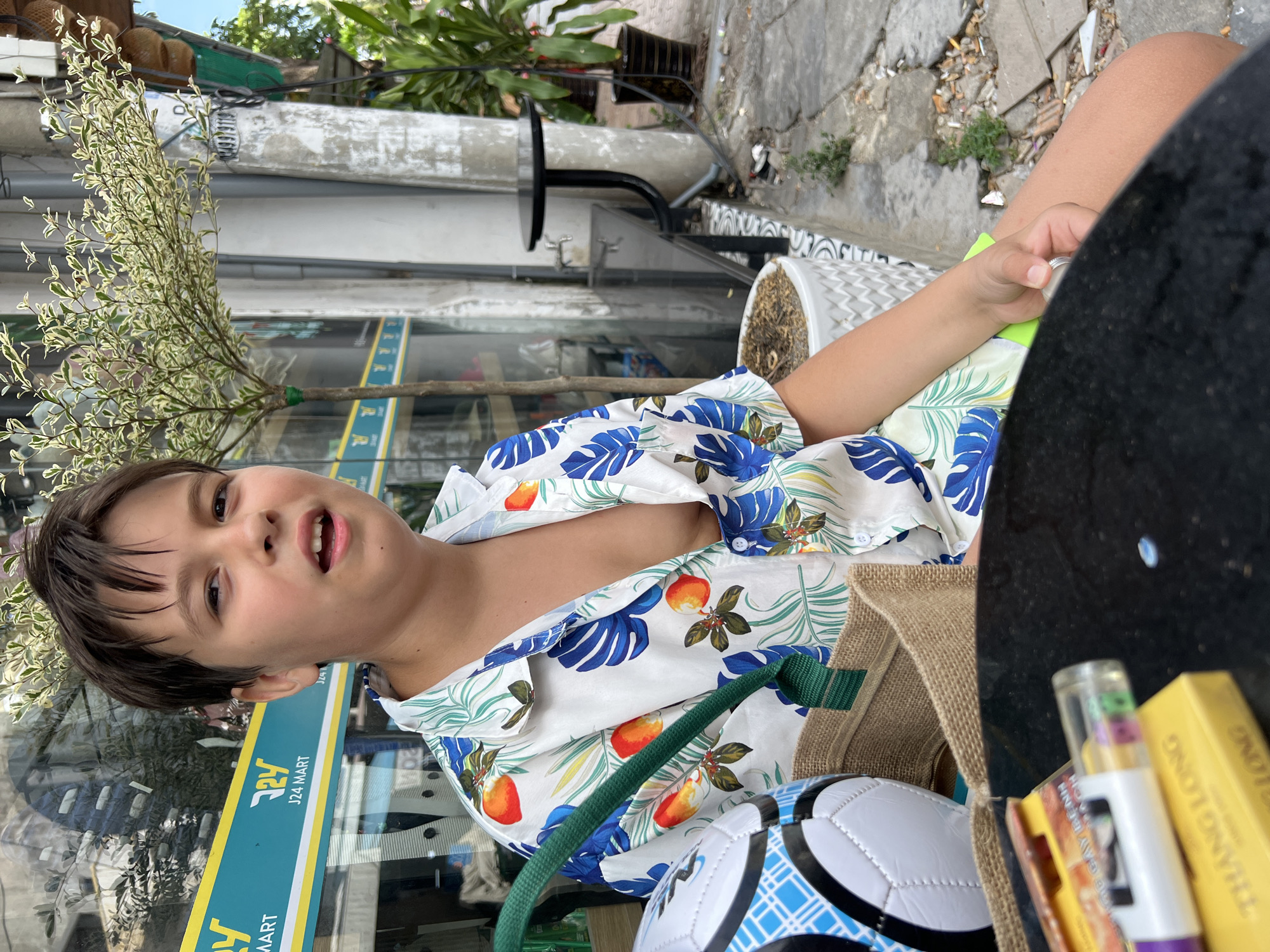 A young Caucasian boy with short dark hair and an open mouth sits in front of a store window in Nha Trang, Vietnam. He is wearing a short-sleeved, collared shirt with a tropical print of oranges and blue leaves, and green shorts. In front of him, a burlap bag contains a white and blue soccer ball. To his left, a black table is partially visible with cigarette packs and a lighter on it. The store window behind him has a yellow and blue sign that reads "J24 MART." A small tree with white-and-green leaves is planted in a white pot to the boy's right. The background shows a concrete sidewalk with plants and a metal table and chairs.