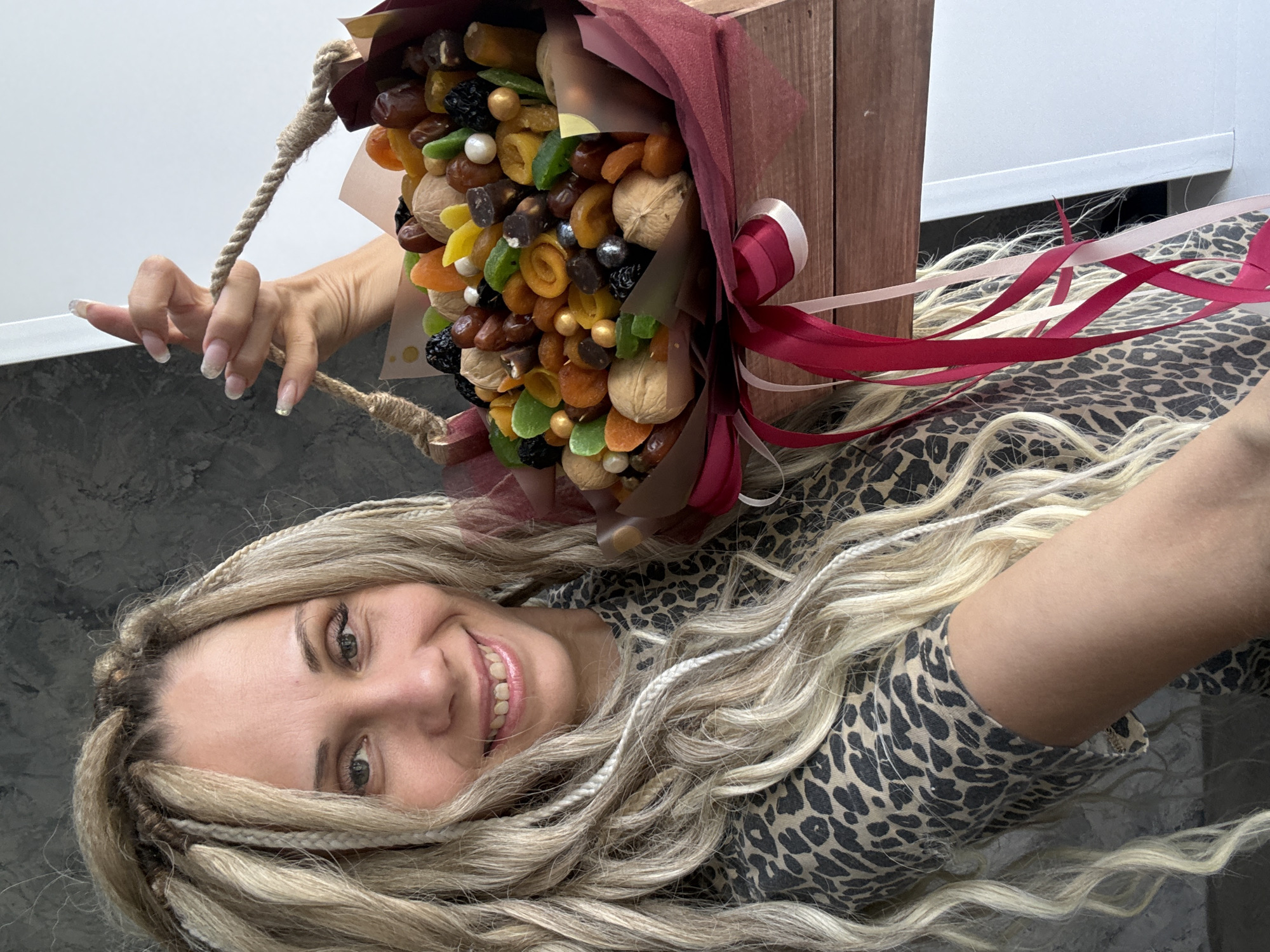 A person with blonde hair, dressed in a leopard-print top, is holding a bouquet of dried fruits and nuts. The bouquet is contained within a wooden box and tied with red and white ribbons. The location appears to be indoors, near a white wall and a gray surface. The city is Unknown, Unknown.