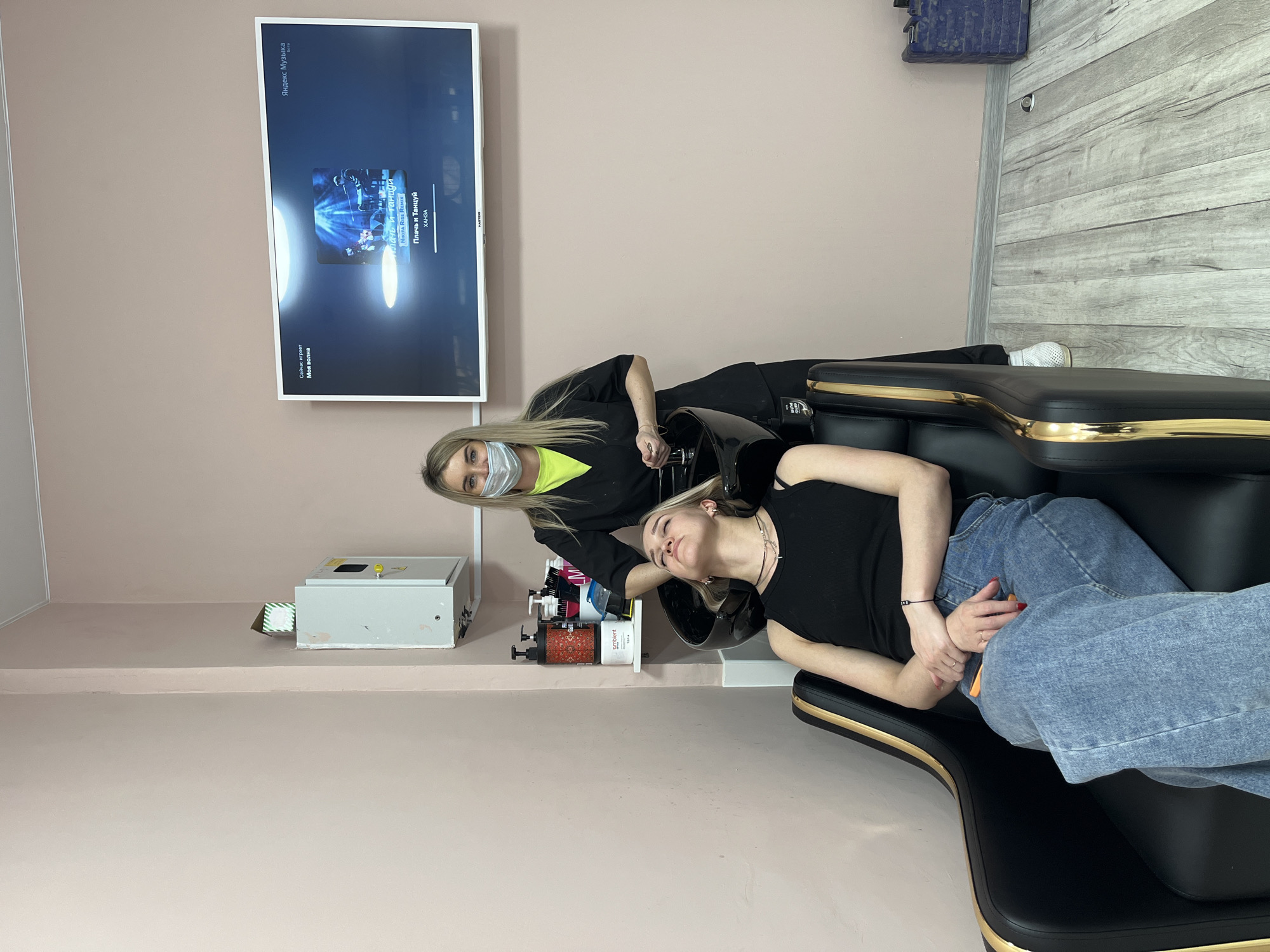 In a brightly lit indoor setting in what appears to be a salon in the city of Chaikovsky, Russia, a woman with blonde hair is reclining in a black, gold-trimmed hair-washing station, with her head positioned in the sink. She is wearing a black tank top and blue jeans, her hands clasped together on her stomach.
Standing behind her is another woman, presumably a hair stylist, dressed in a black top with a bright yellow undershirt and a face mask. Her blonde hair is pulled back. She is in the process of washing or rinsing the client's hair, her hands near the sink area.
Behind the stylist, mounted on the light pink wall, is a TV screen displaying music information, specifically indicating that "Плачь и Танцуй" by ХАНЗА is playing on Яндекс Музыка. A metal electrical box is visible on a shelf to the left. Several bottles of hair products are arranged near the sink. The floor is a light wood laminate.