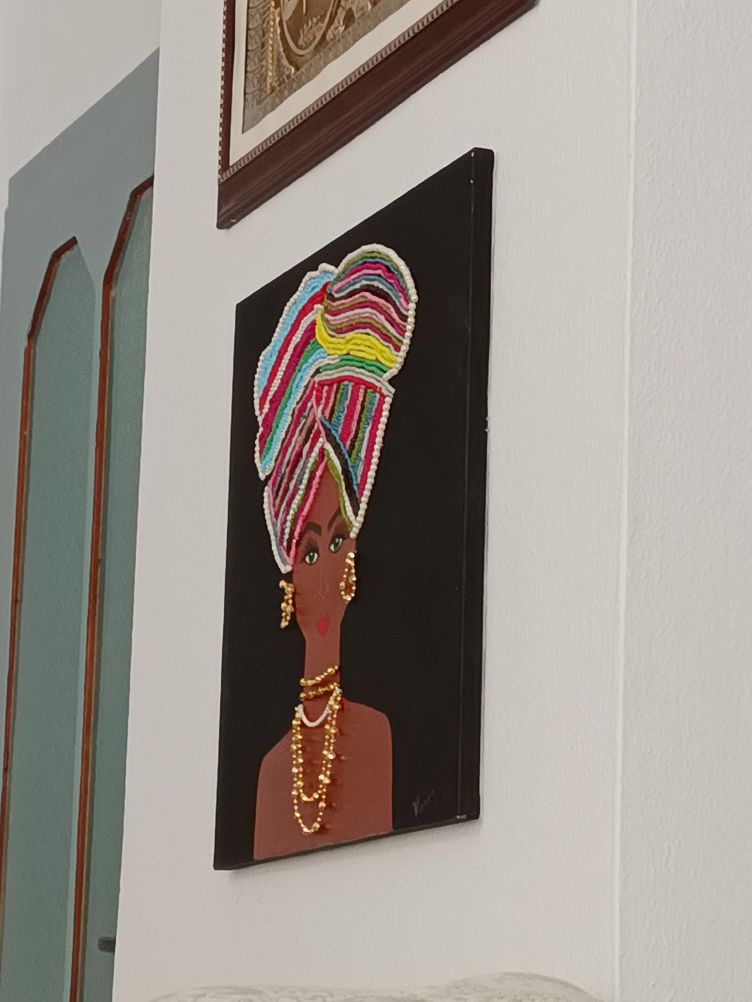 The image shows a framed painting of a Black woman wearing a colorful headwrap and jewelry. The painting is displayed on a white wall. To the left of the painting, there is a doorway or archway with a blue-green color. Above the painting, another framed artwork is partially visible. The scene appears to be an indoor setting. The painting is the main subject, with the woman depicted in a stylized manner, featuring bright, multi-colored stripes on her headwrap and gold beads for her necklace and earrings. Her skin tone is brown, and she has green eyes and red lips. The background of the painting is black. The overall impression is one of vibrant decoration. The location is specified as Alanya, Turkey, though there are no specific visual cues in the image to confirm this geographical location beyond the provided prompt.